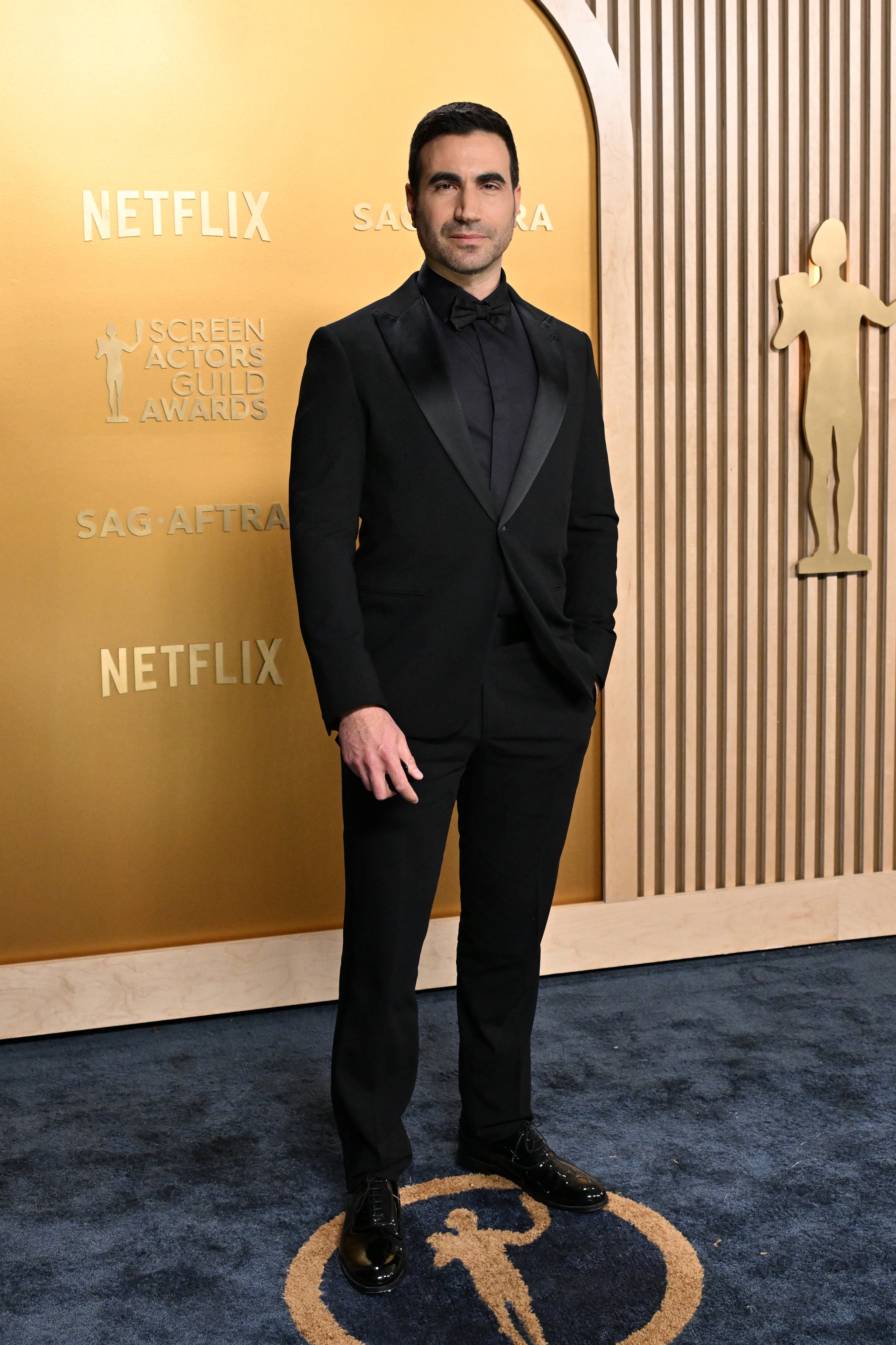 Person in a tailored suit stands confidently on a red carpet at an awards event backdrop