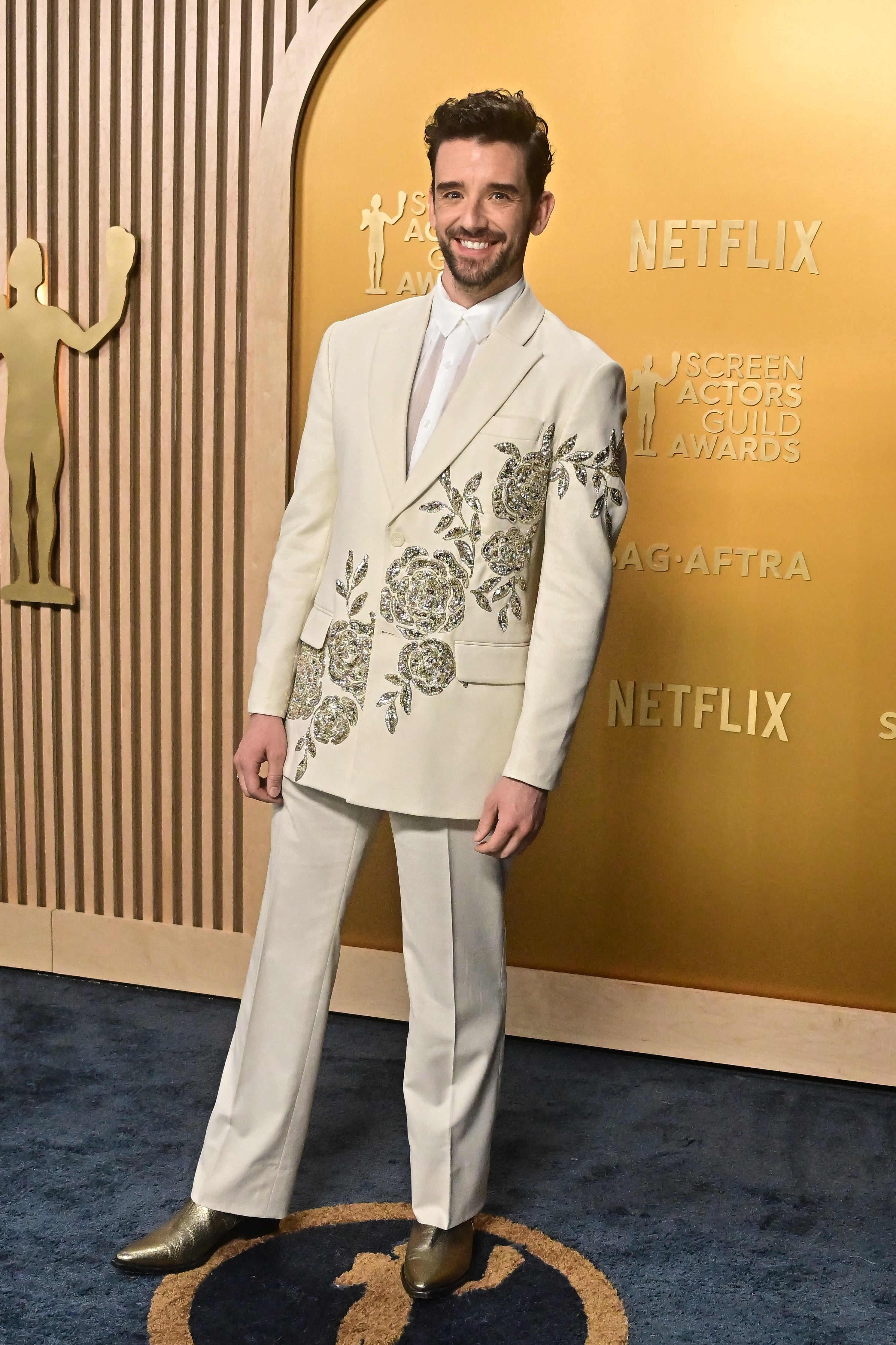 Person in embroidered suit and tie on a red carpet at an awards event, smiling. Backdrop displays event logos