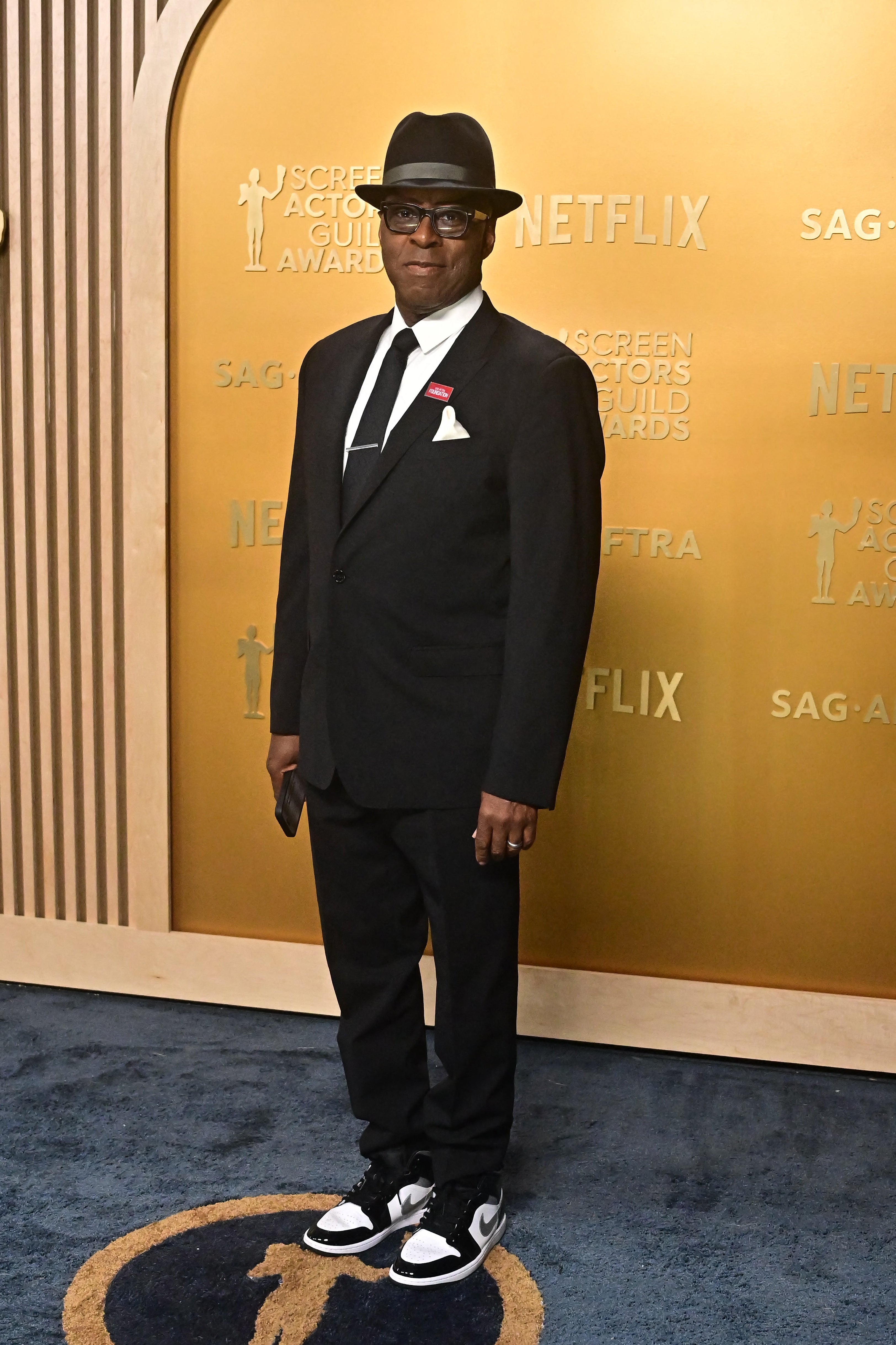 Person in a black suit, white tie, fedora, and sneakers on a red carpet at a SAG event, standing against a background with event signage