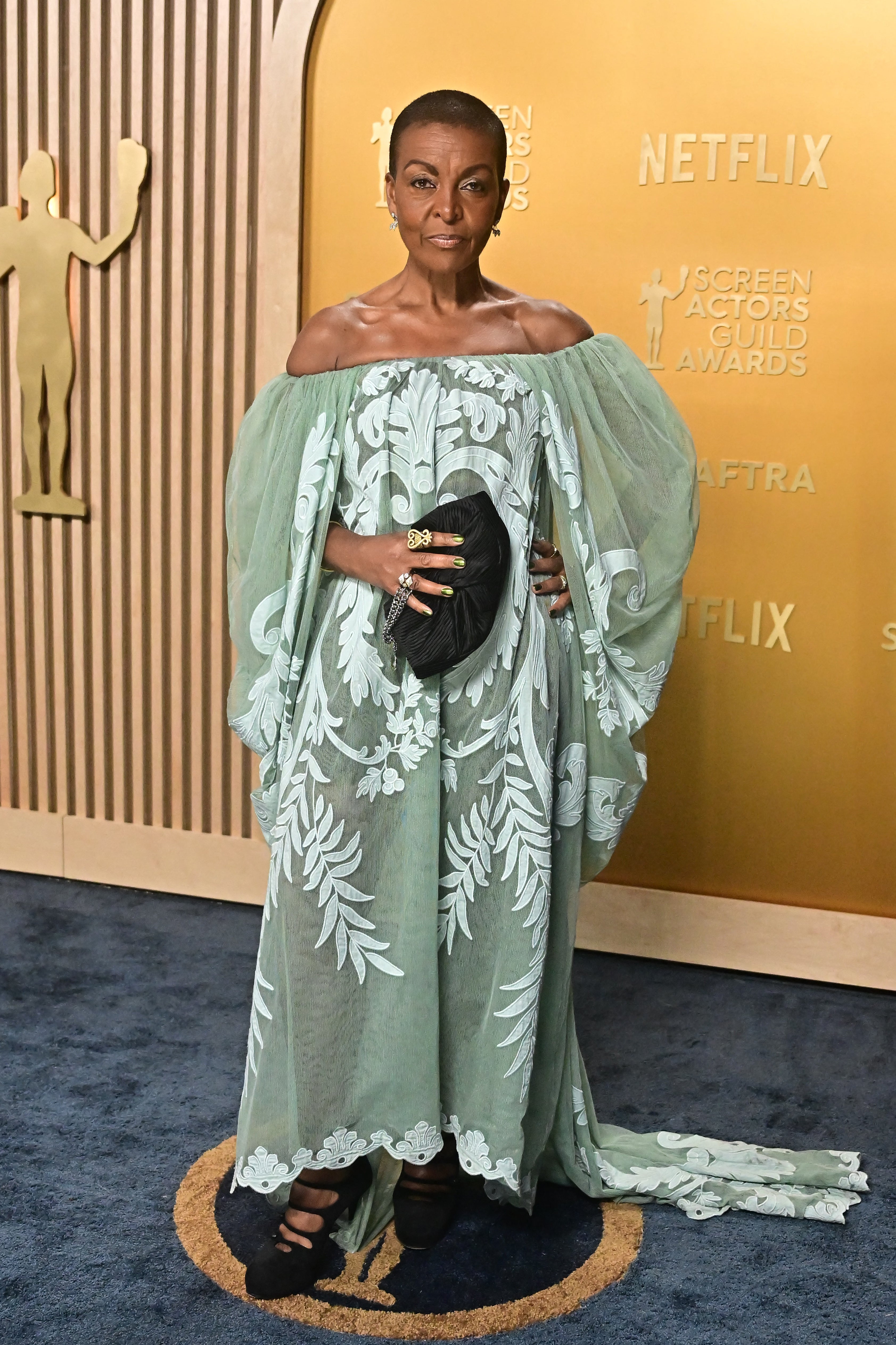 Person in an elegant off-the-shoulder dress with floral patterns, holding a clutch. Background has "Screen Actors Guild Awards" signage