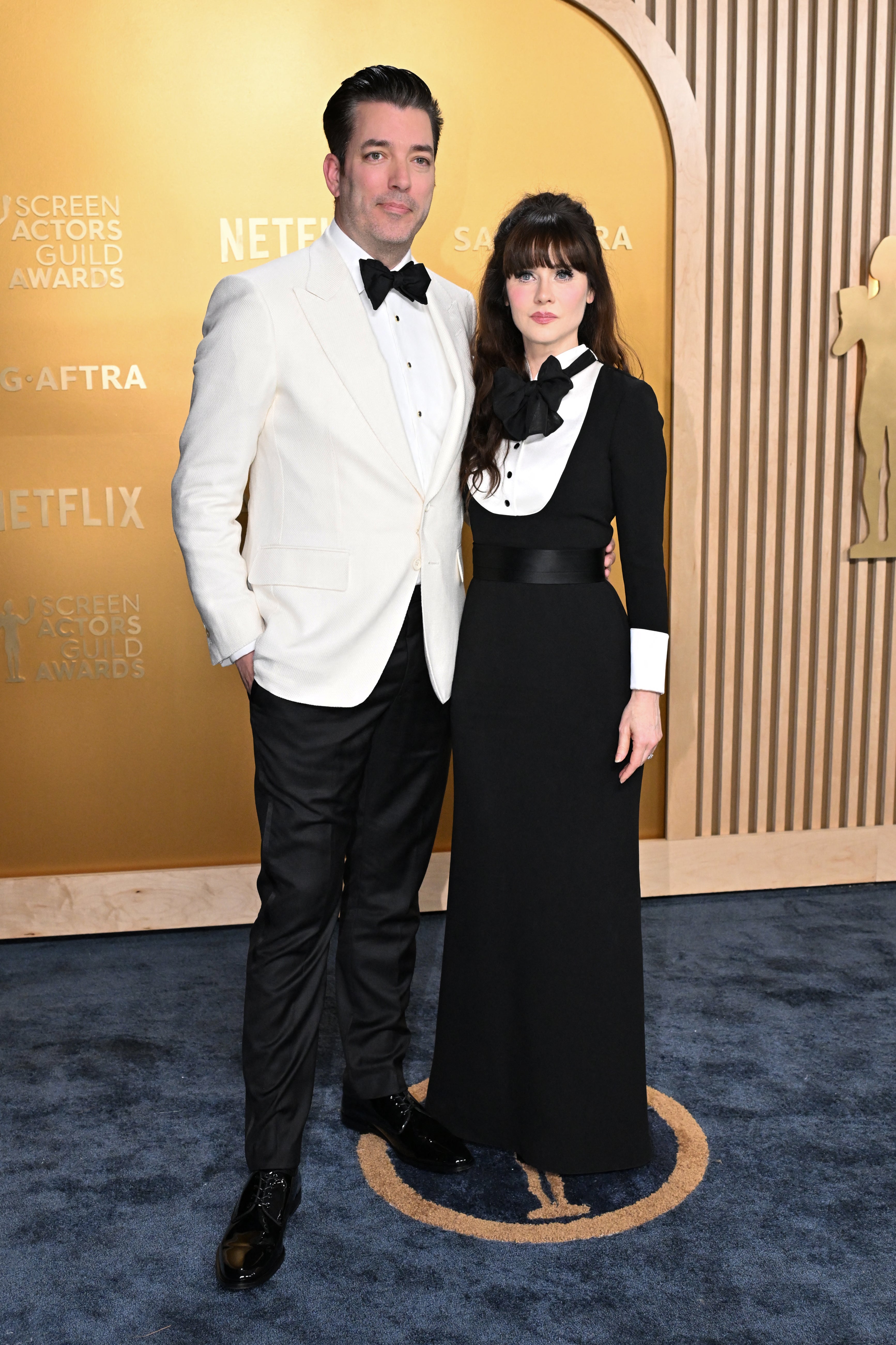 A couple poses on a red carpet. The man wears a white tuxedo jacket and black pants; the woman wears a long black dress with white accents
