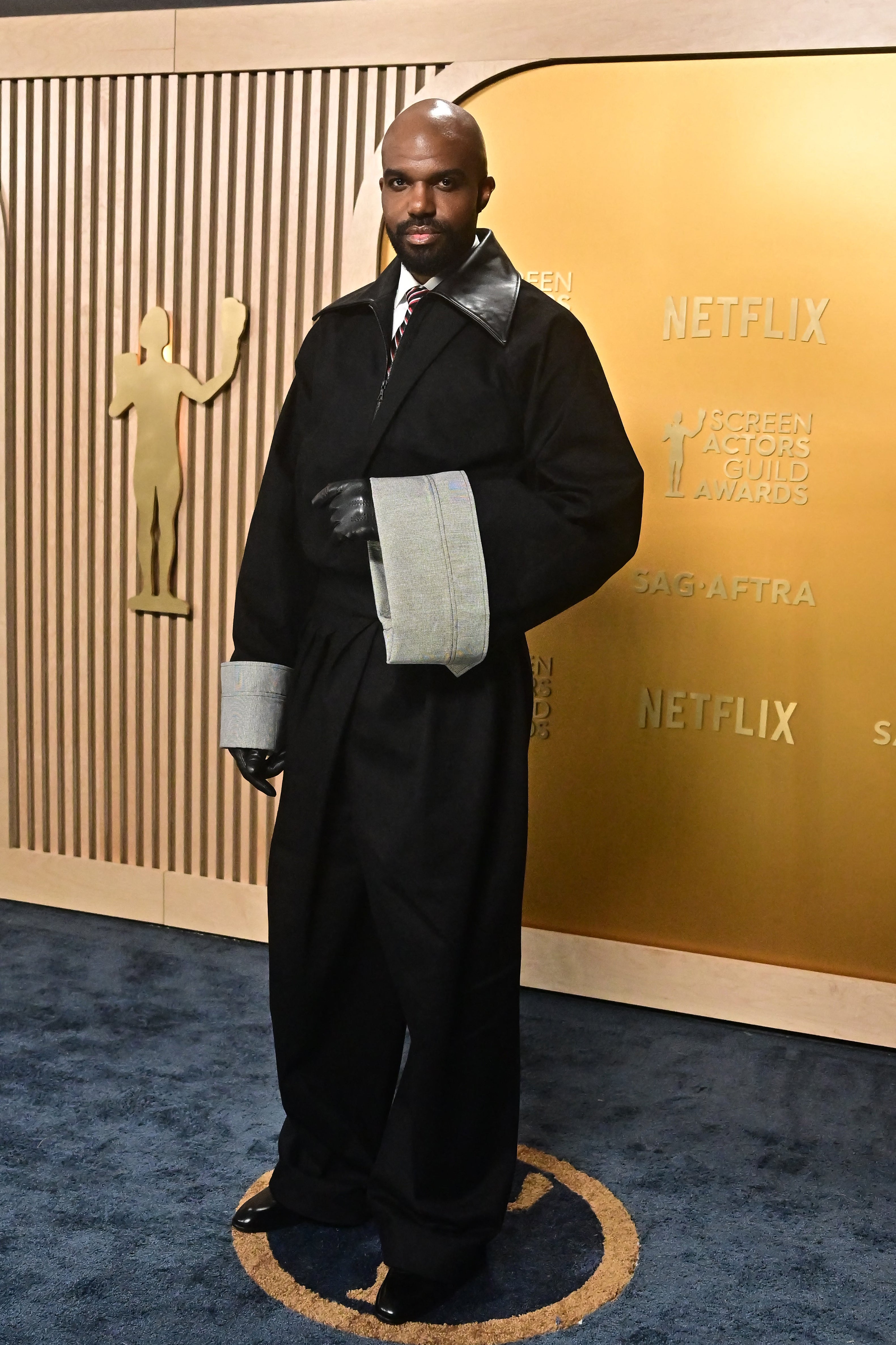 Person on red carpet in formal attire with oversized cuffs, posing for photos at an event with Netflix and SAG-AFTRA logos in the background