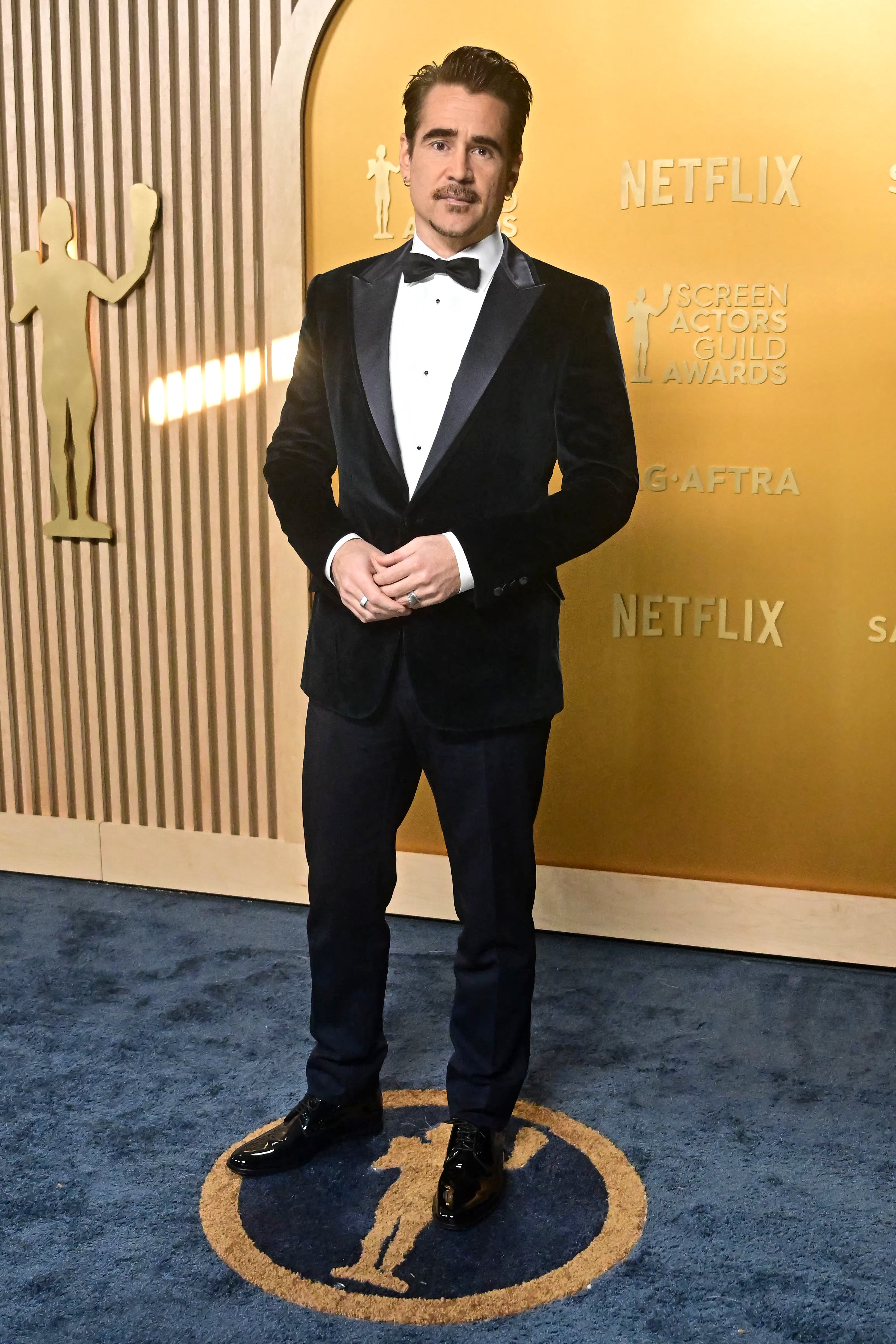 Man in a tuxedo stands on a blue carpet at an awards event, with a backdrop featuring logos and symbols