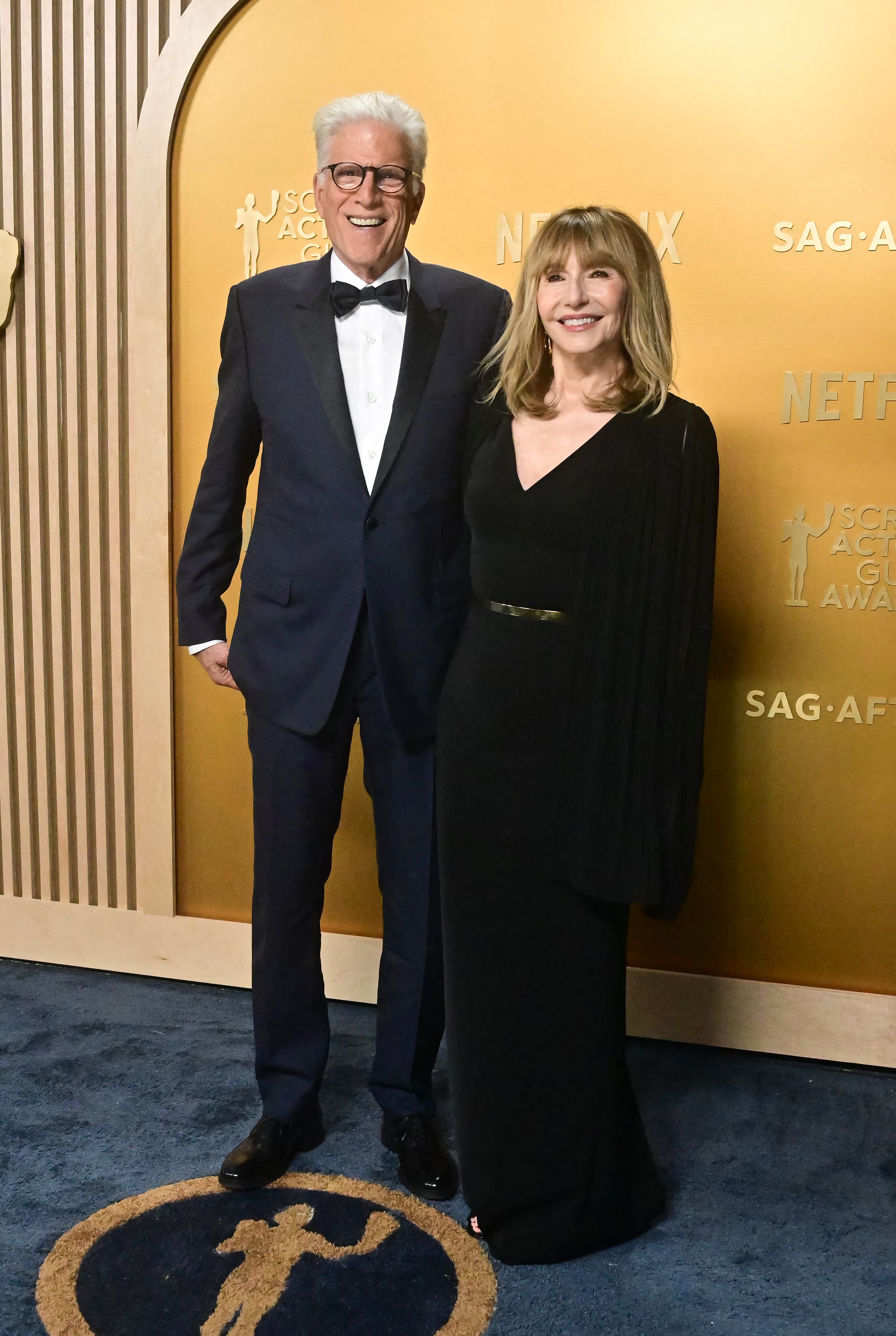 Two people on a red carpet, one in a tuxedo and the other in a sleek, long-sleeved gown, posing at a formal event