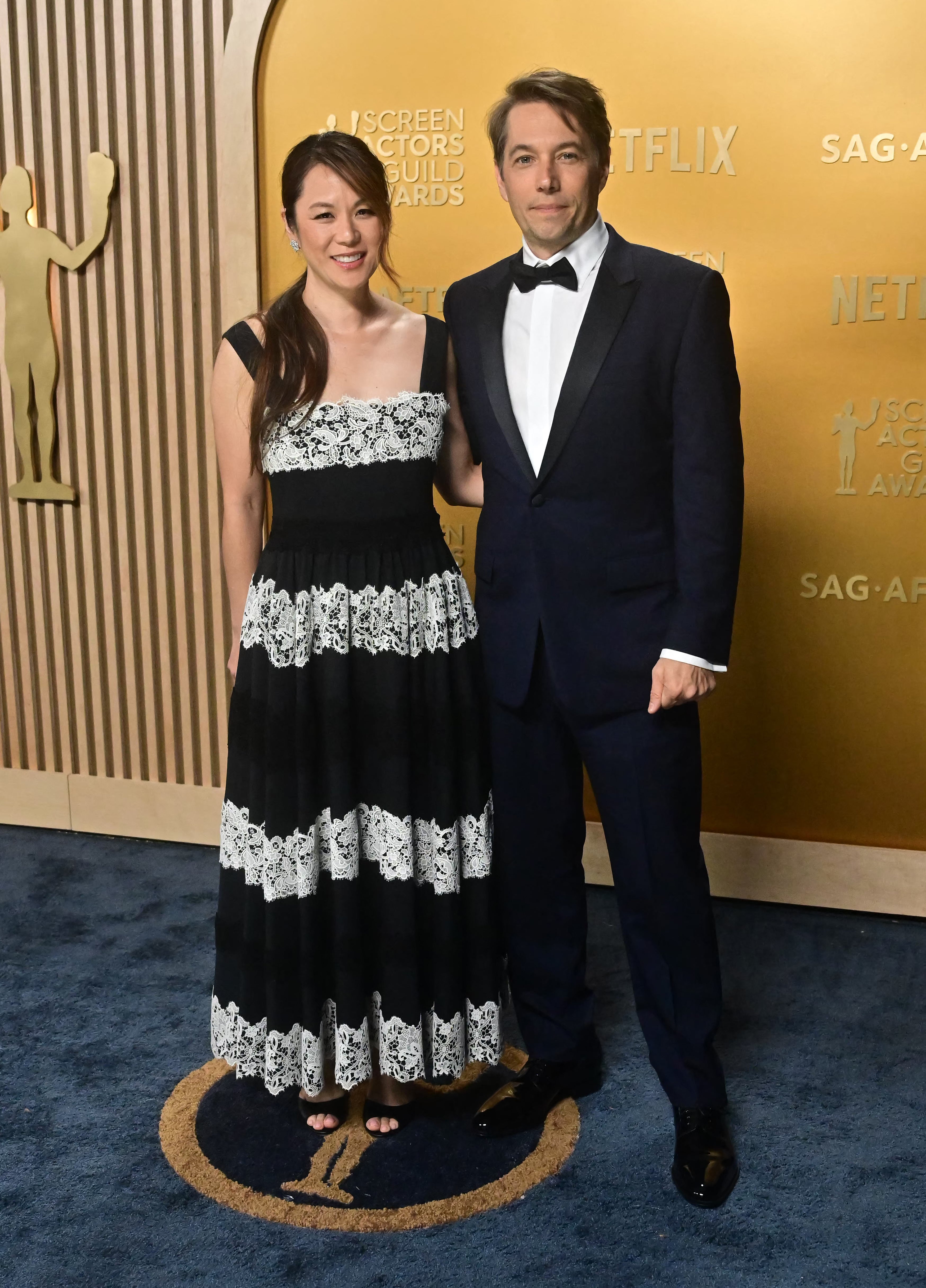 Two people on a red carpet. The woman wears a black and white lace dress; the man is in a classic tuxedo