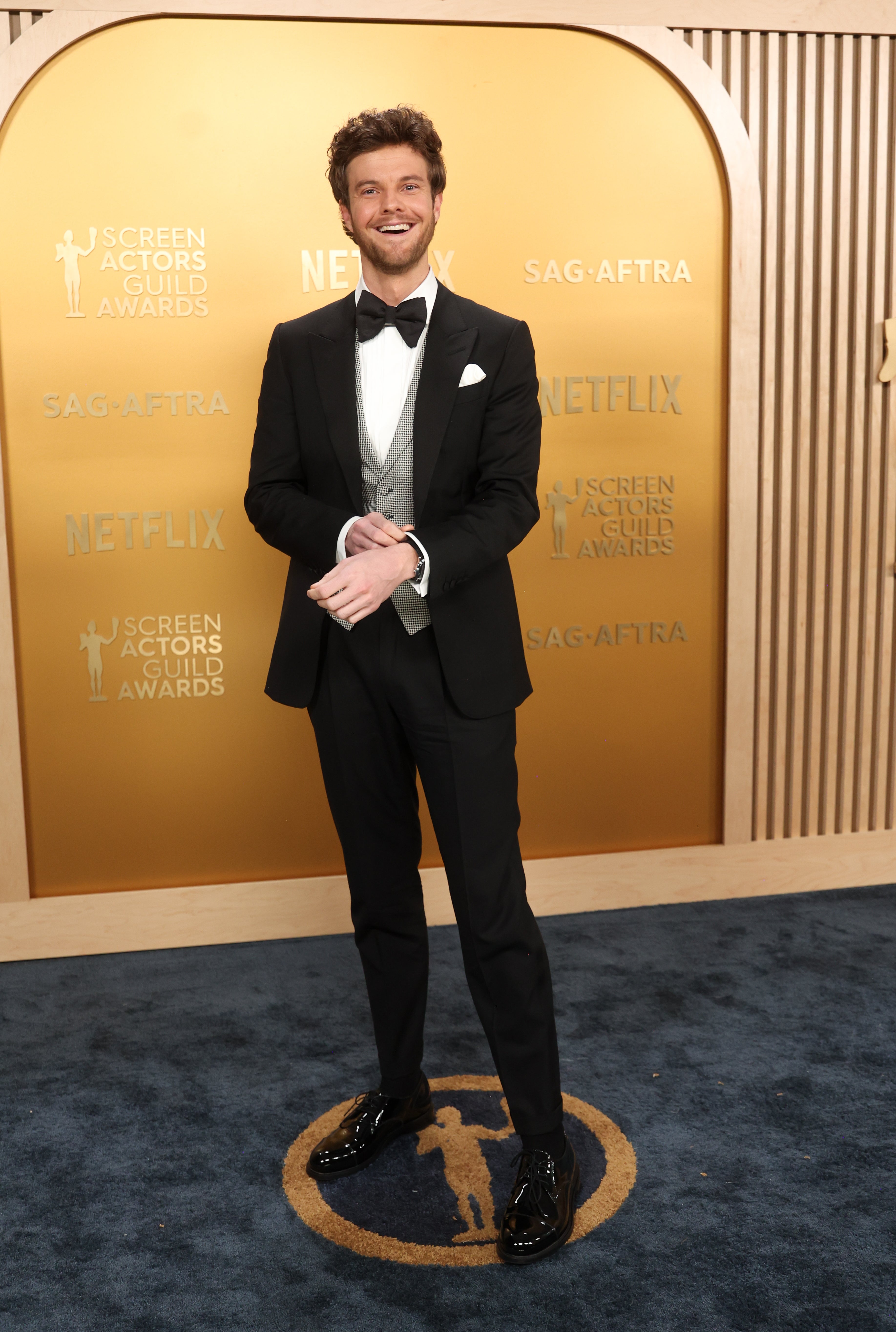 Person in formal suit with bow tie stands on a red carpet at an award event backdrop
