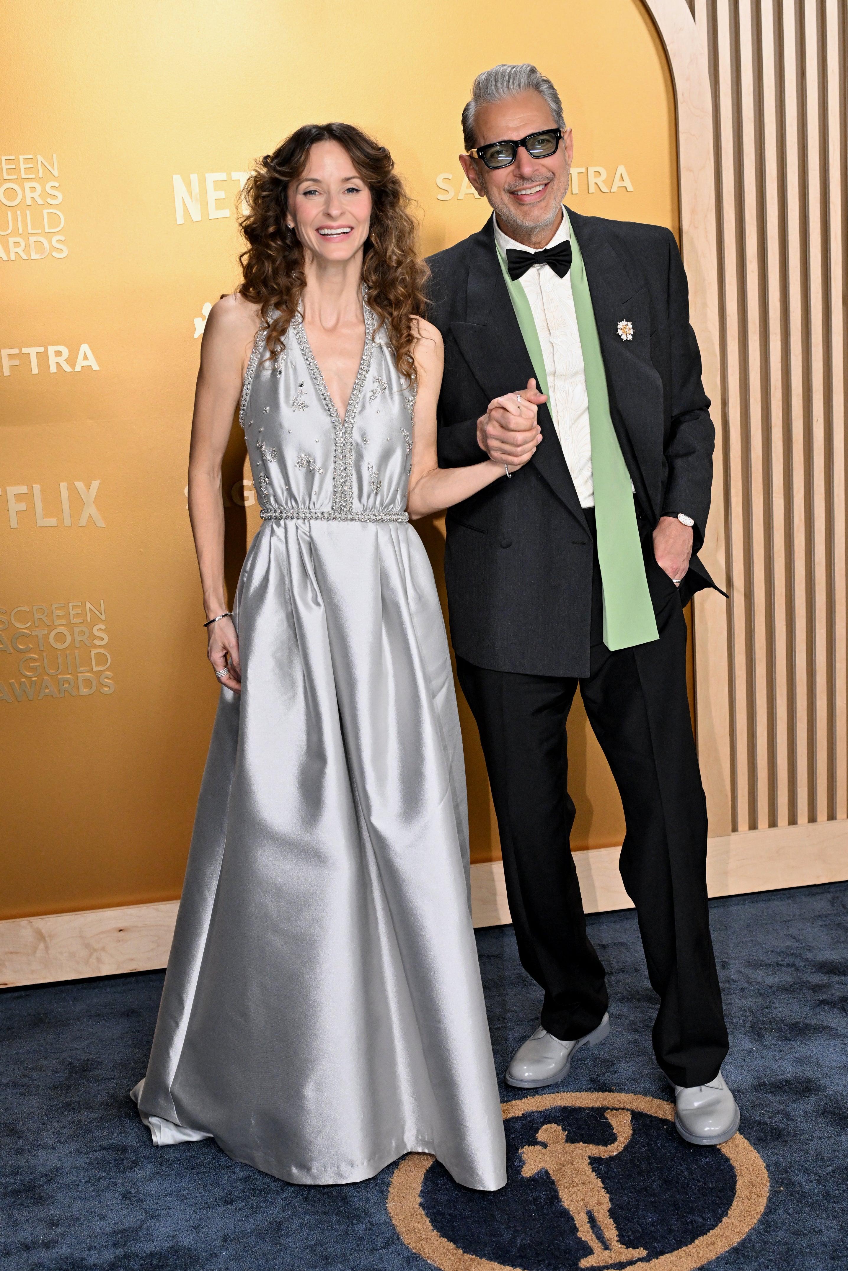 Two people pose on a red carpet. One wears an elegant long gown, and the other wears a suit with a green tie and sunglasses, holding hands