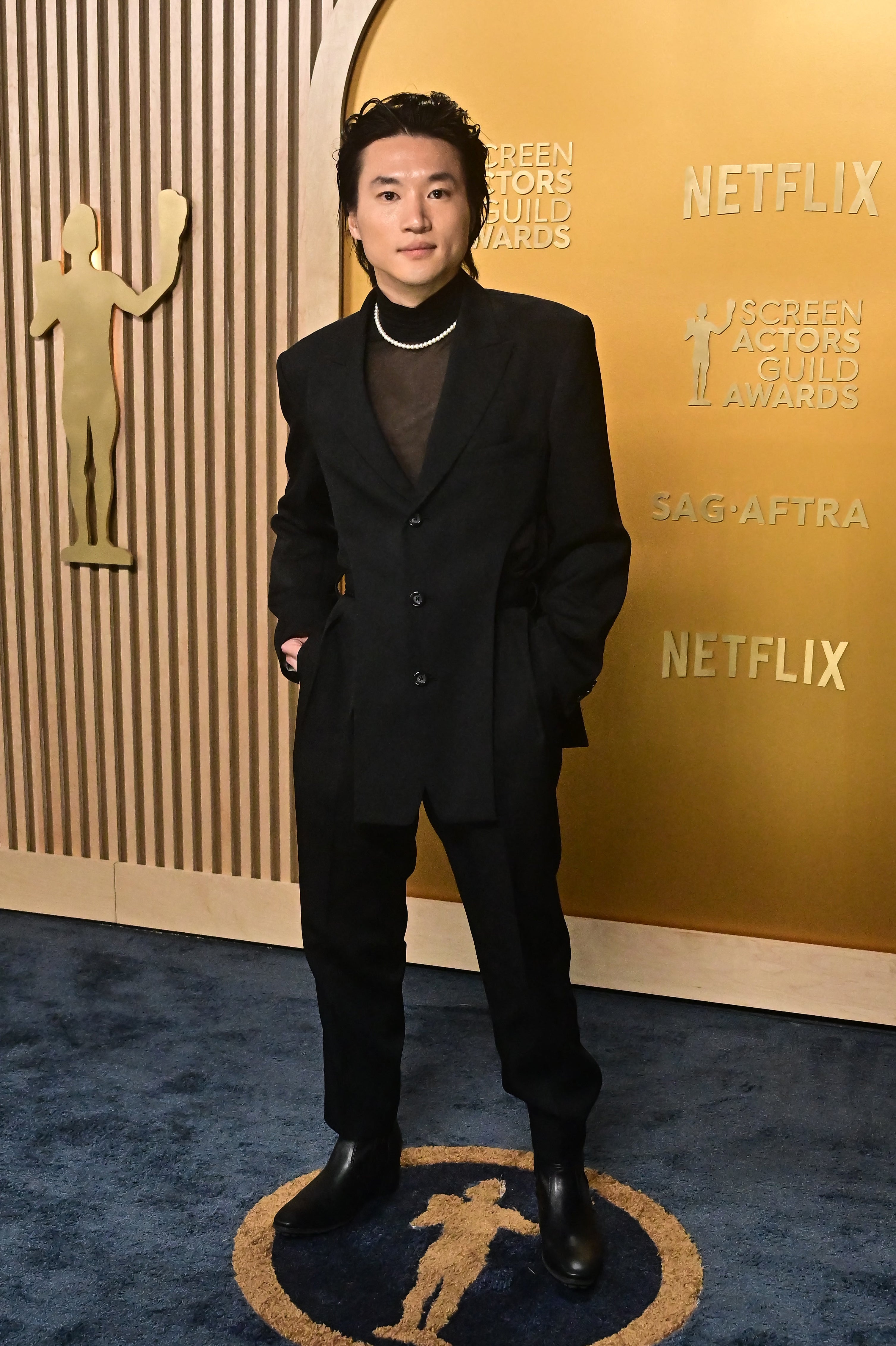 Person in a formal suit with a modern style poses on an awards event backdrop featuring logos