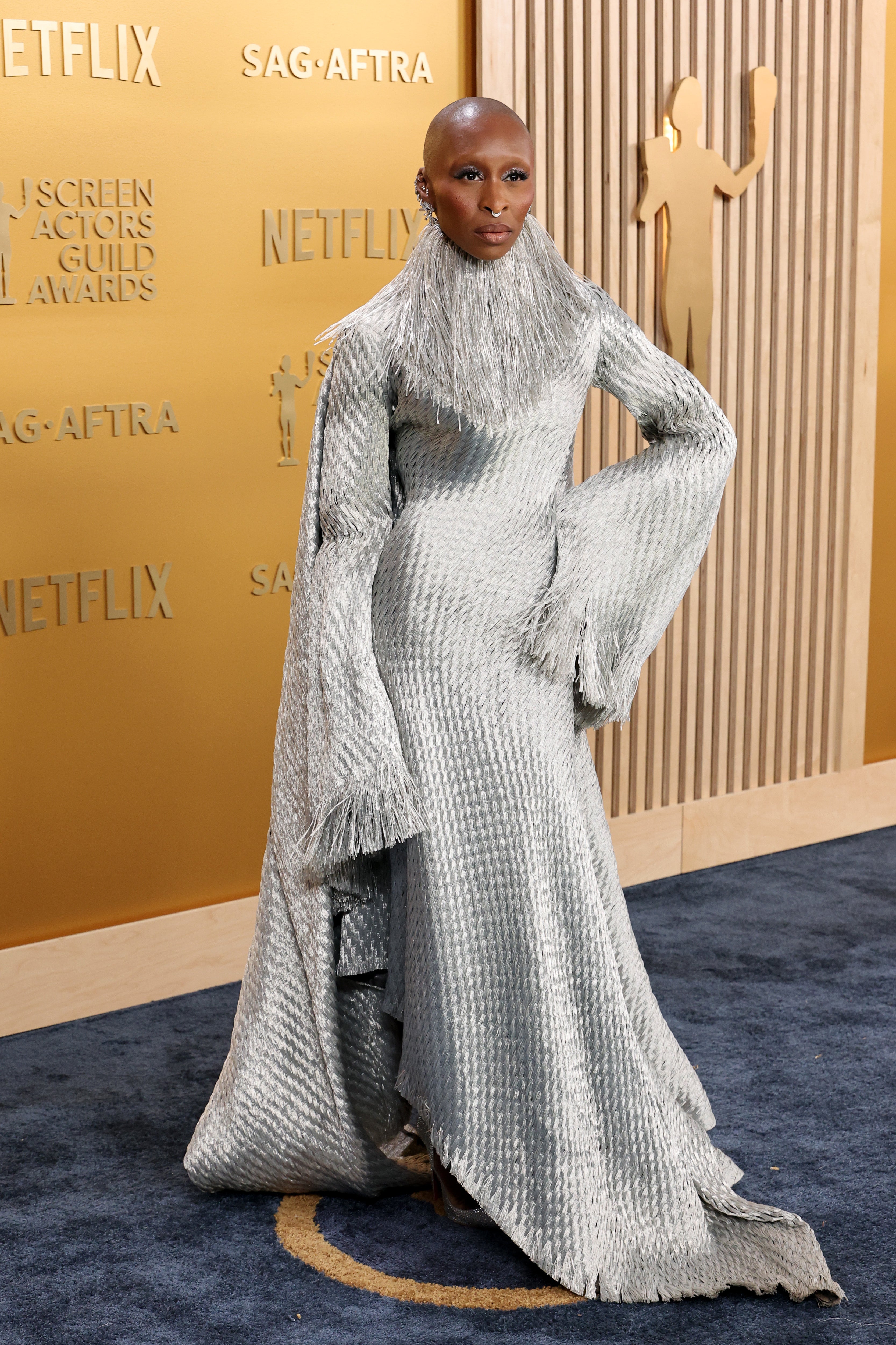 Person in textured, high-collared gown with long sleeves poses at an event backdrop with logos, showcasing a distinctive fashion style