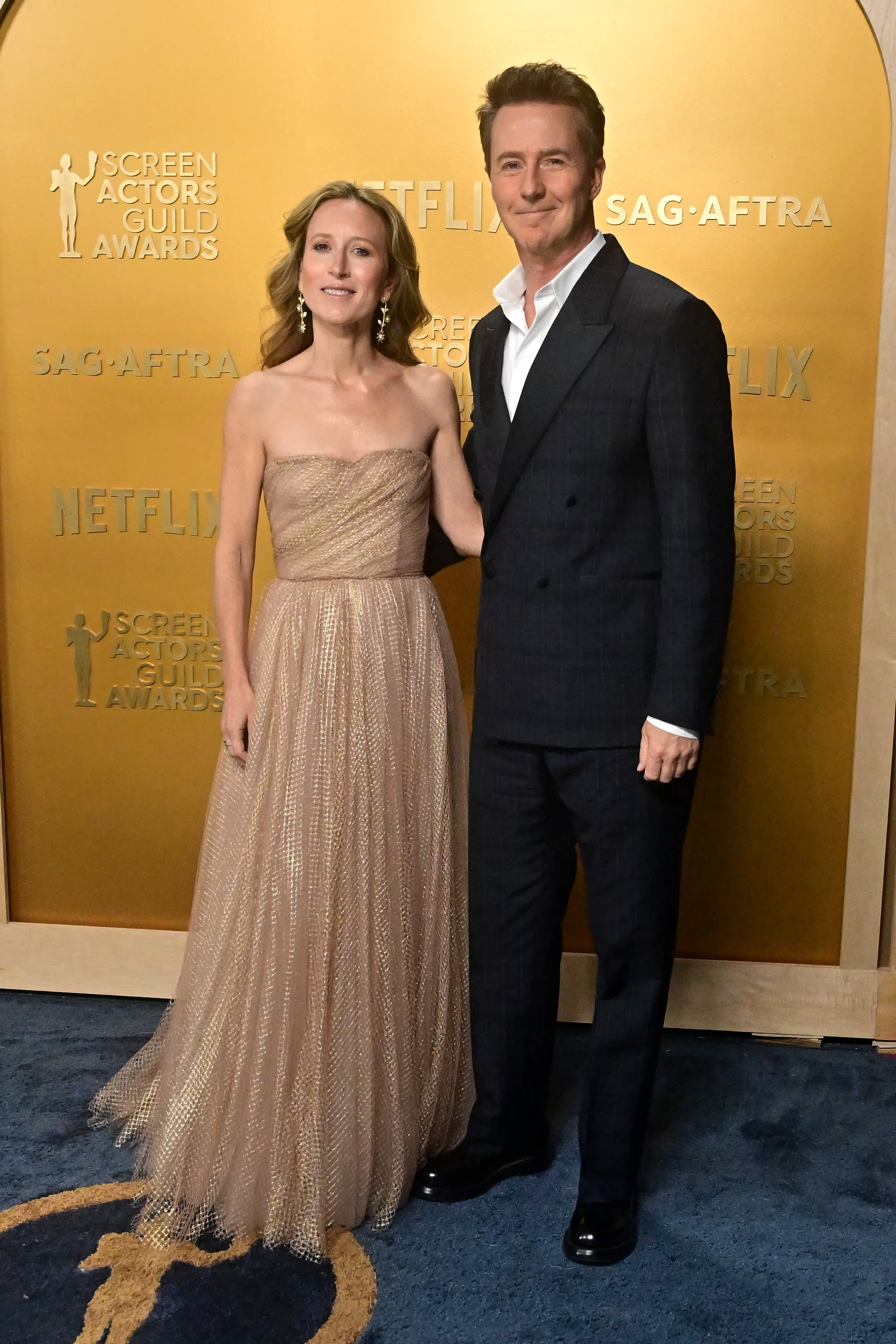 Two individuals in elegant evening attire pose at a Screen Actors Guild Awards event. The woman wears a strapless gown; the man is in a dark suit