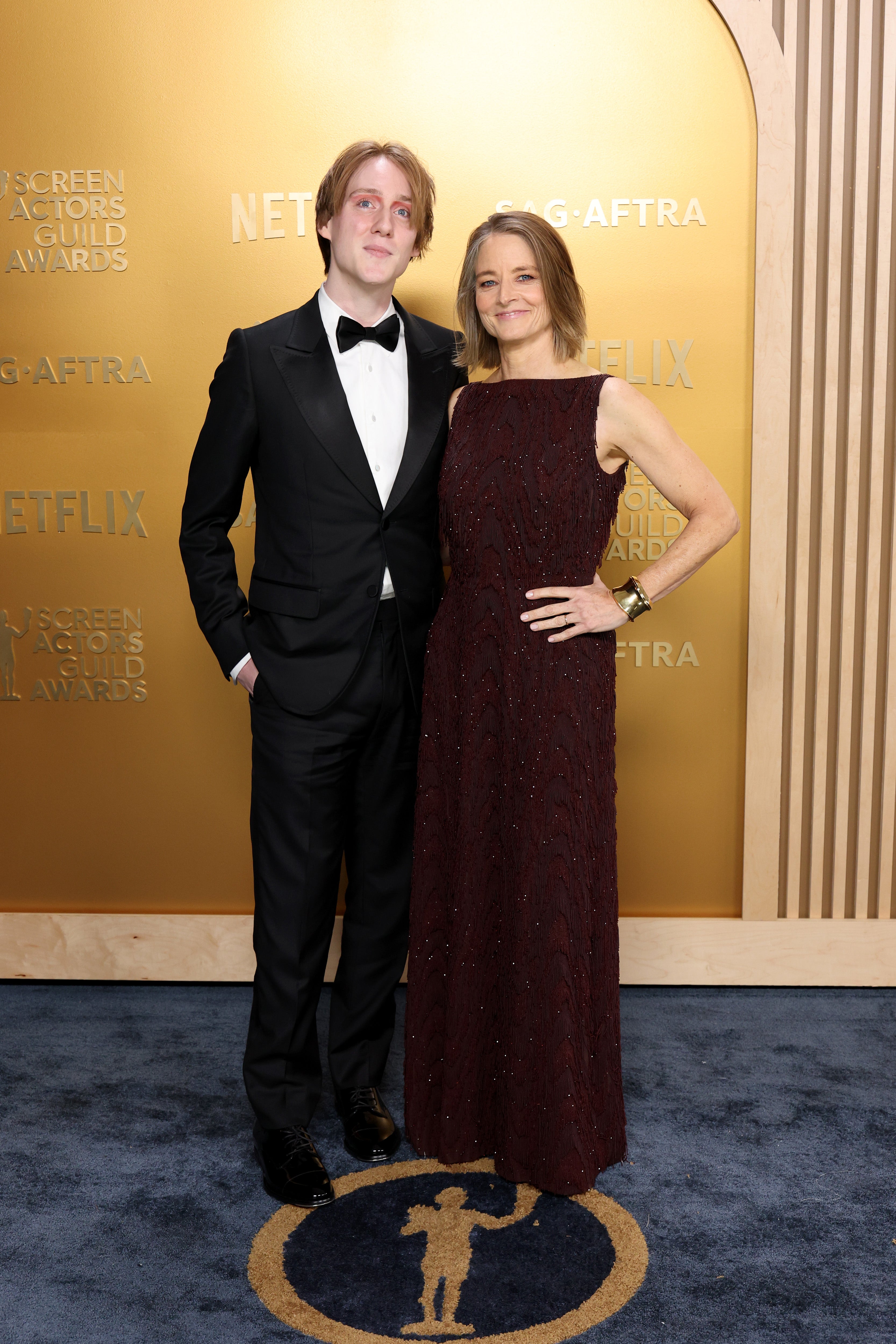 Two people on a red carpet. One wearing a black tuxedo, the other in a sleeveless gown. Logo and "Screen Actors Guild Awards" on the backdrop