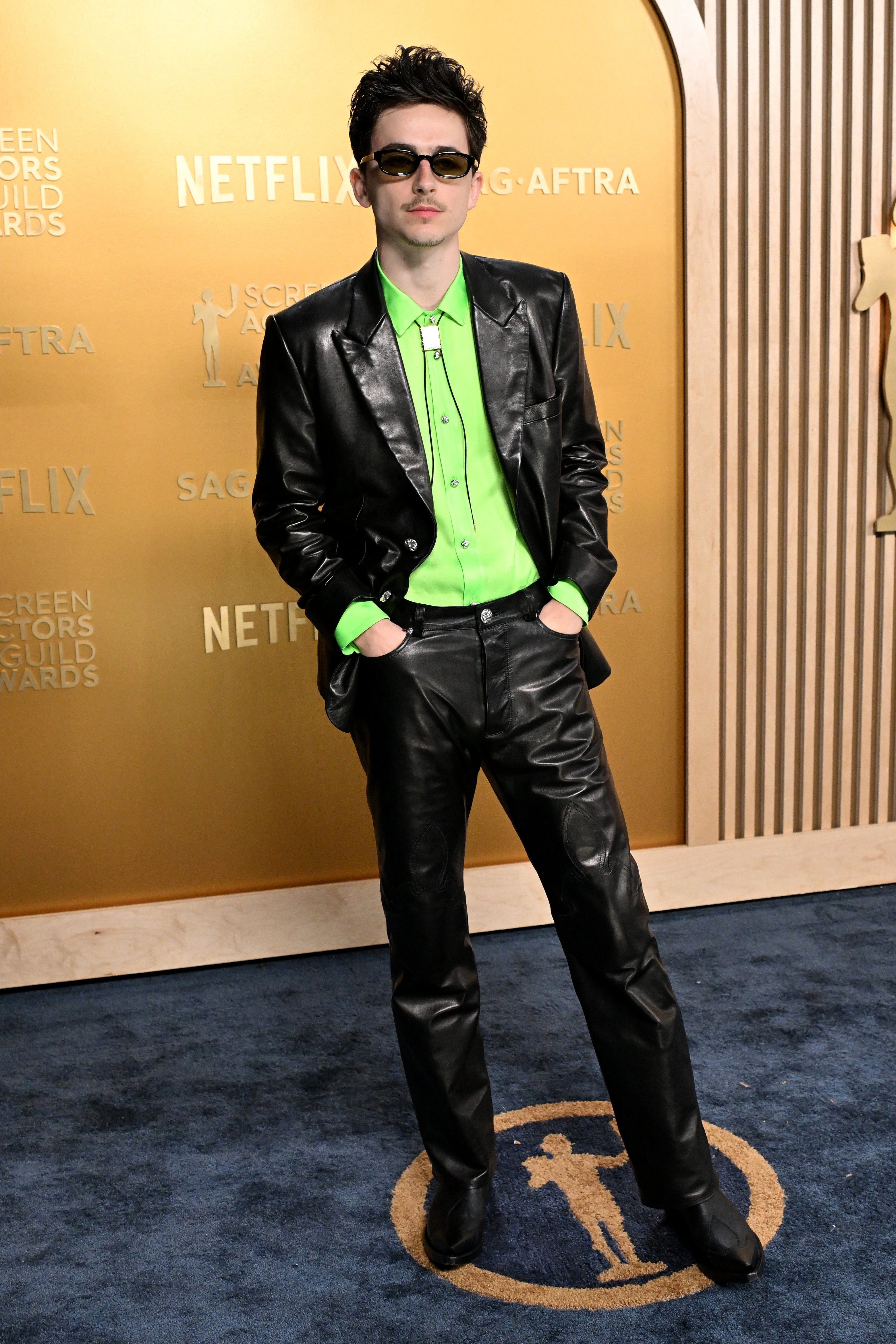 Person in a black leather suit with a neon green shirt and sunglasses on a red carpet with 'Netflix' signage in the background