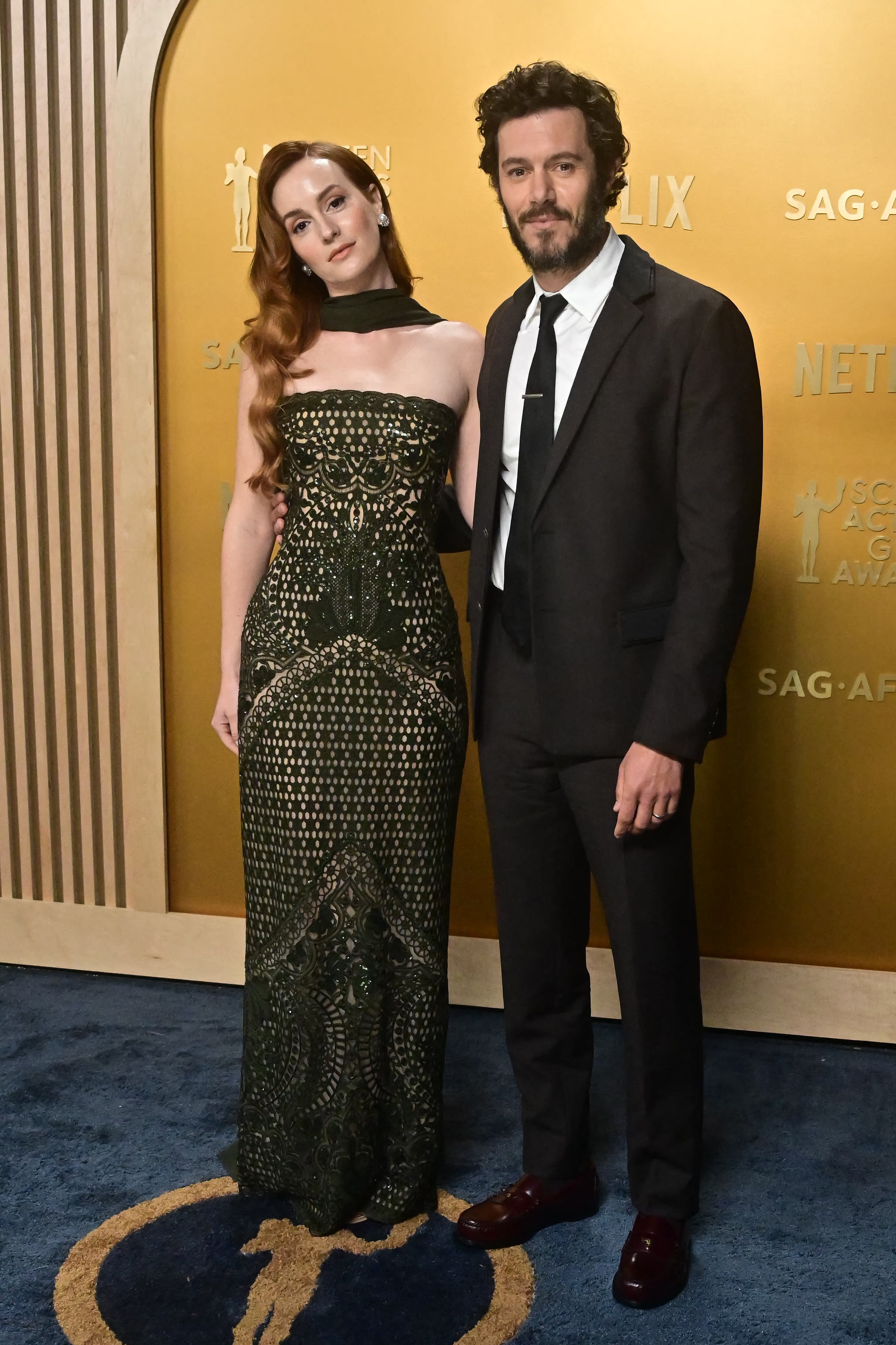 Two individuals pose on a red carpet. The woman wears a patterned strapless gown, and the man is in a suit and tie