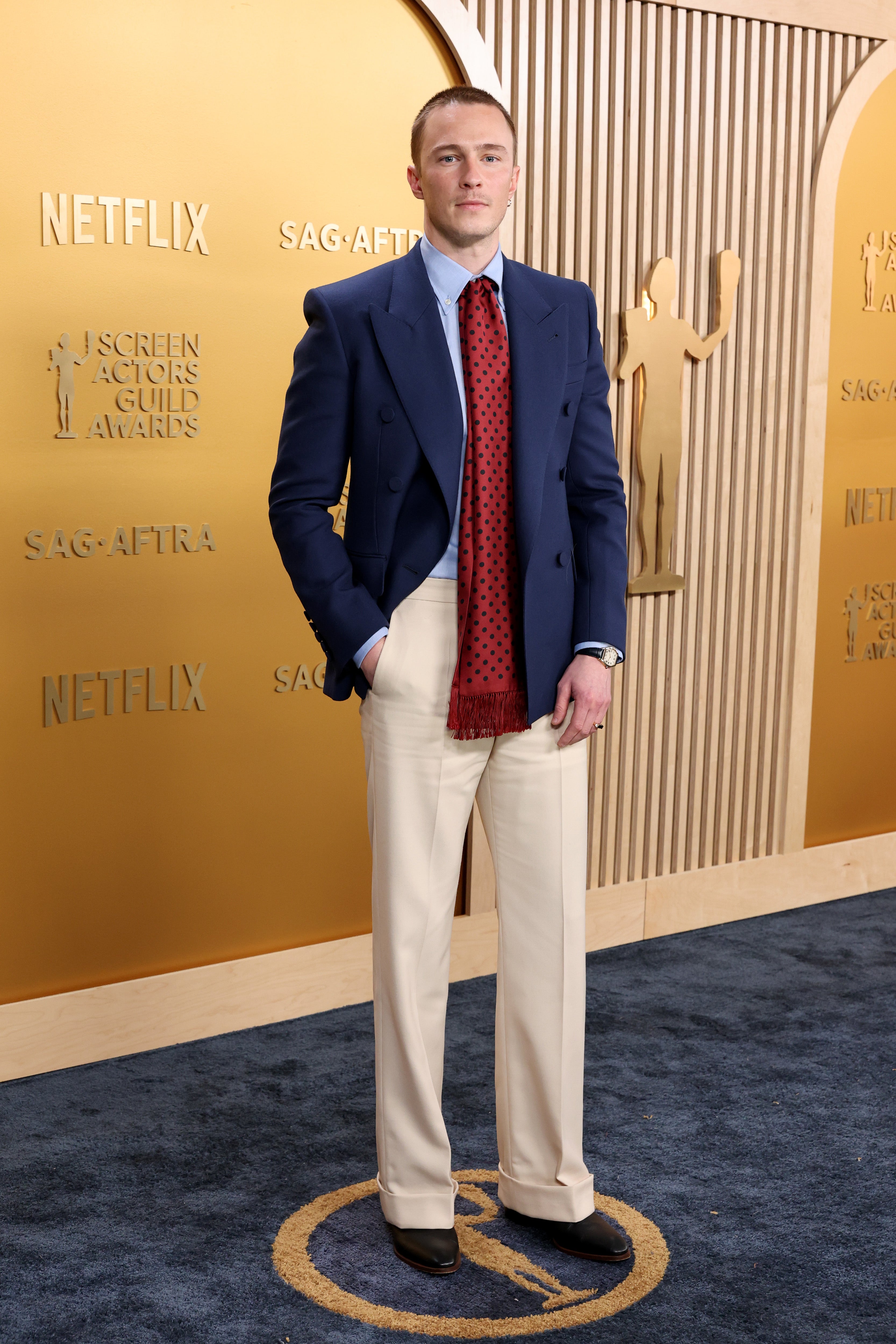 Person on the red carpet wearing a tailored blue blazer with a red polka dot tie and light trousers, standing against a backdrop with award logos
