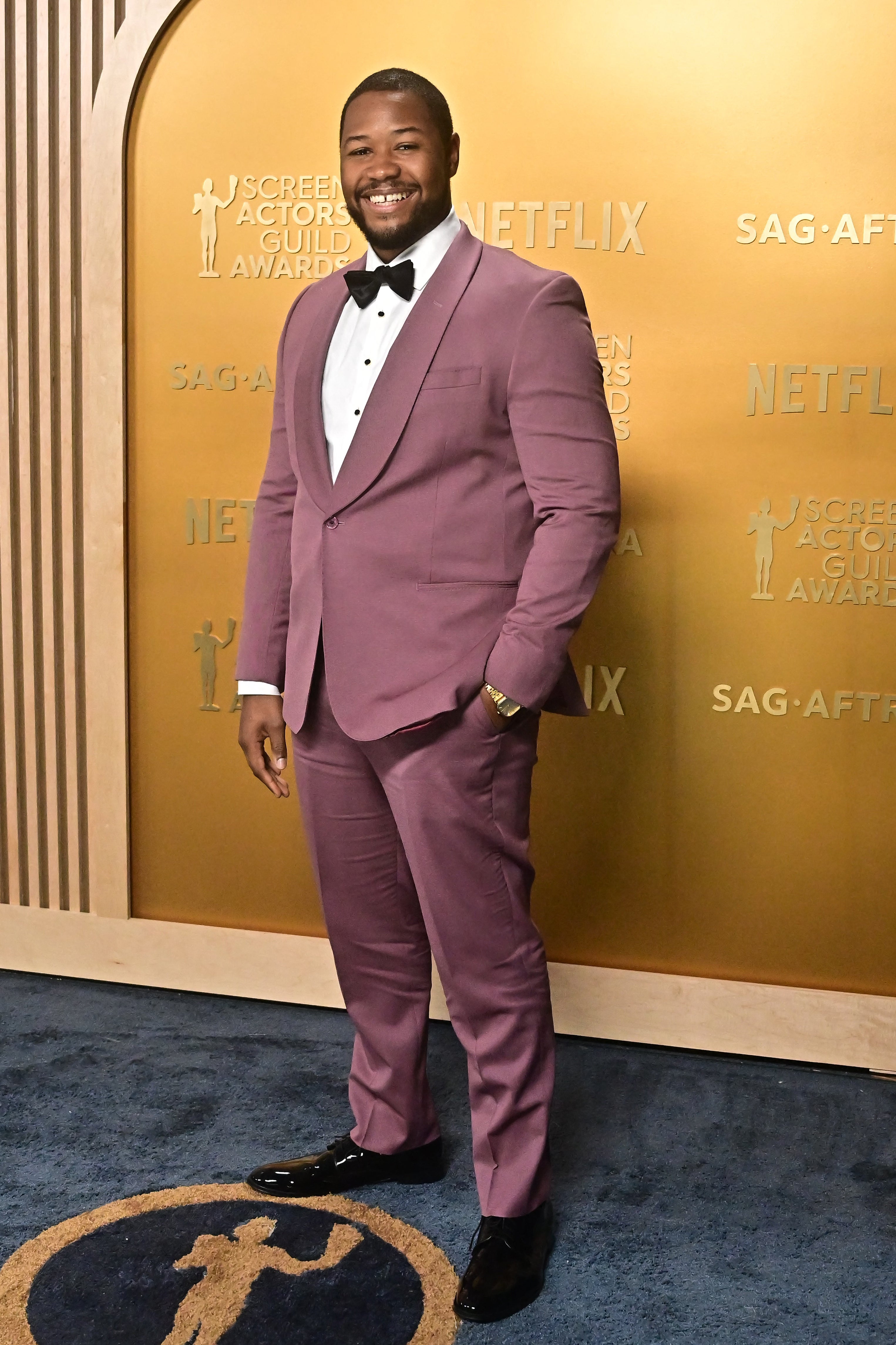 Person in a tailored suit with a white shirt and bow tie standing on a red carpet at an awards event