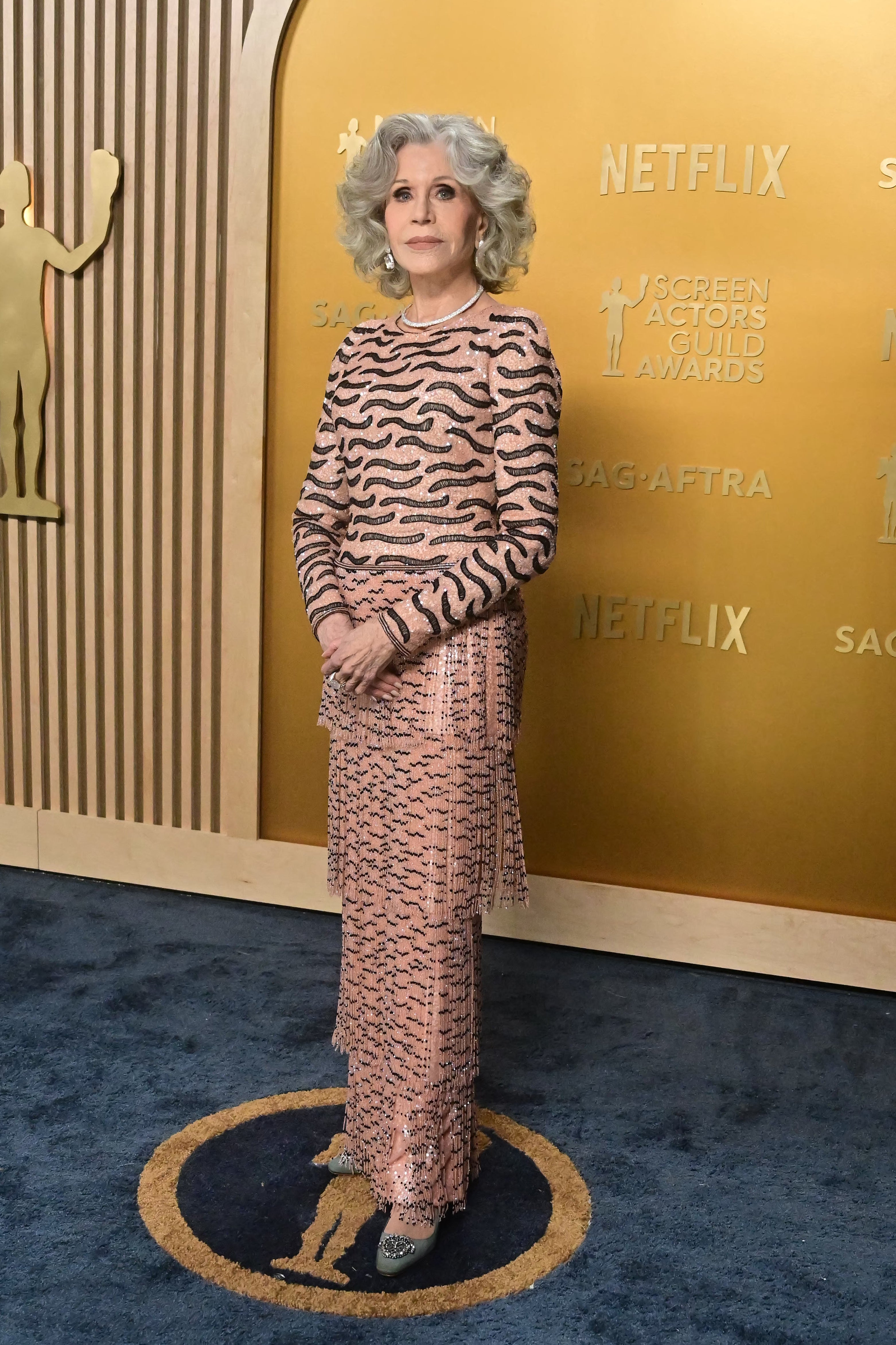 Person on a red carpet wearing a patterned gown with long fringes and intricate details