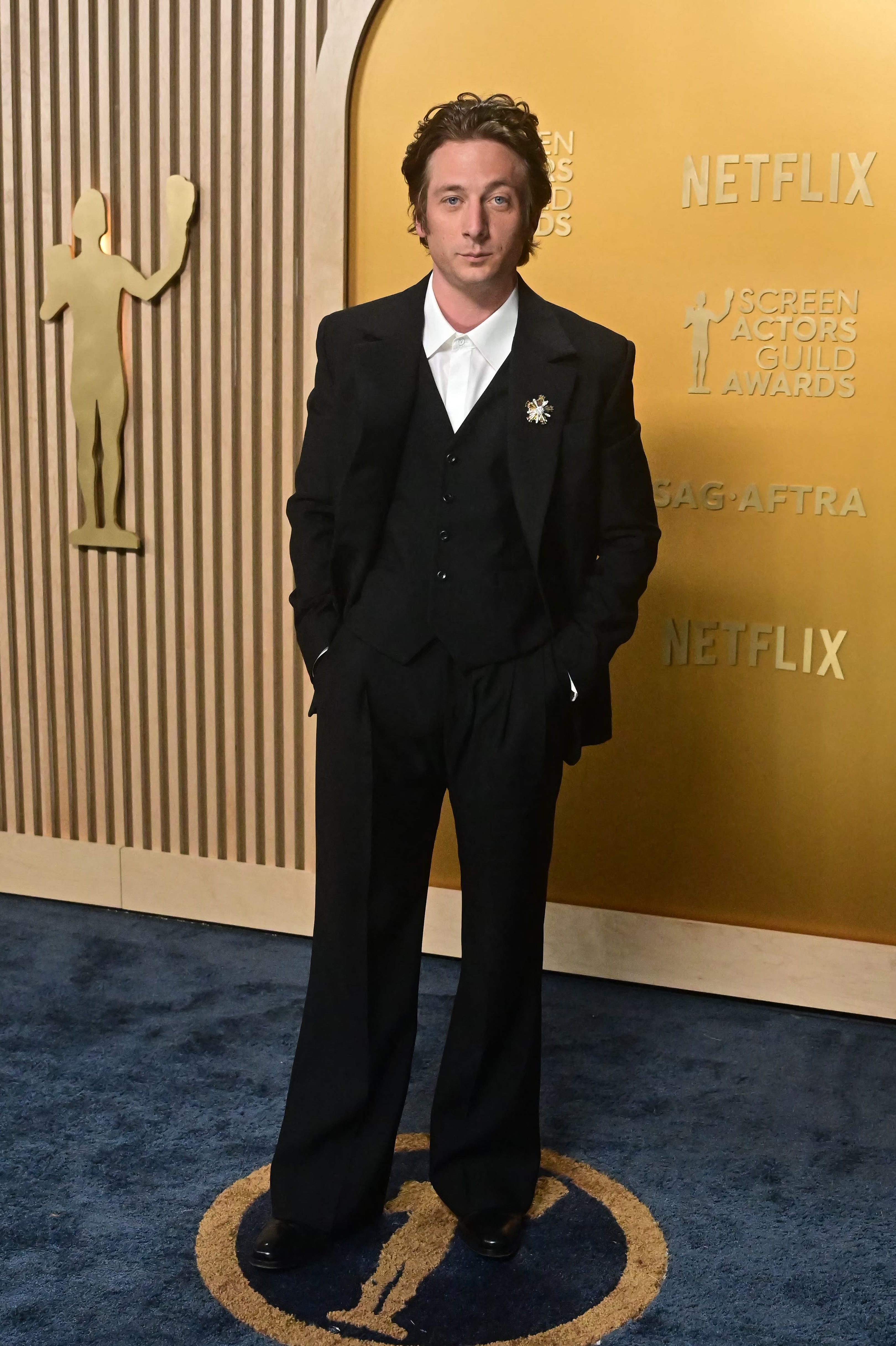 Person in a tailored black suit with wide-leg pants, standing on a blue carpet at an awards event with stylish, classic decor