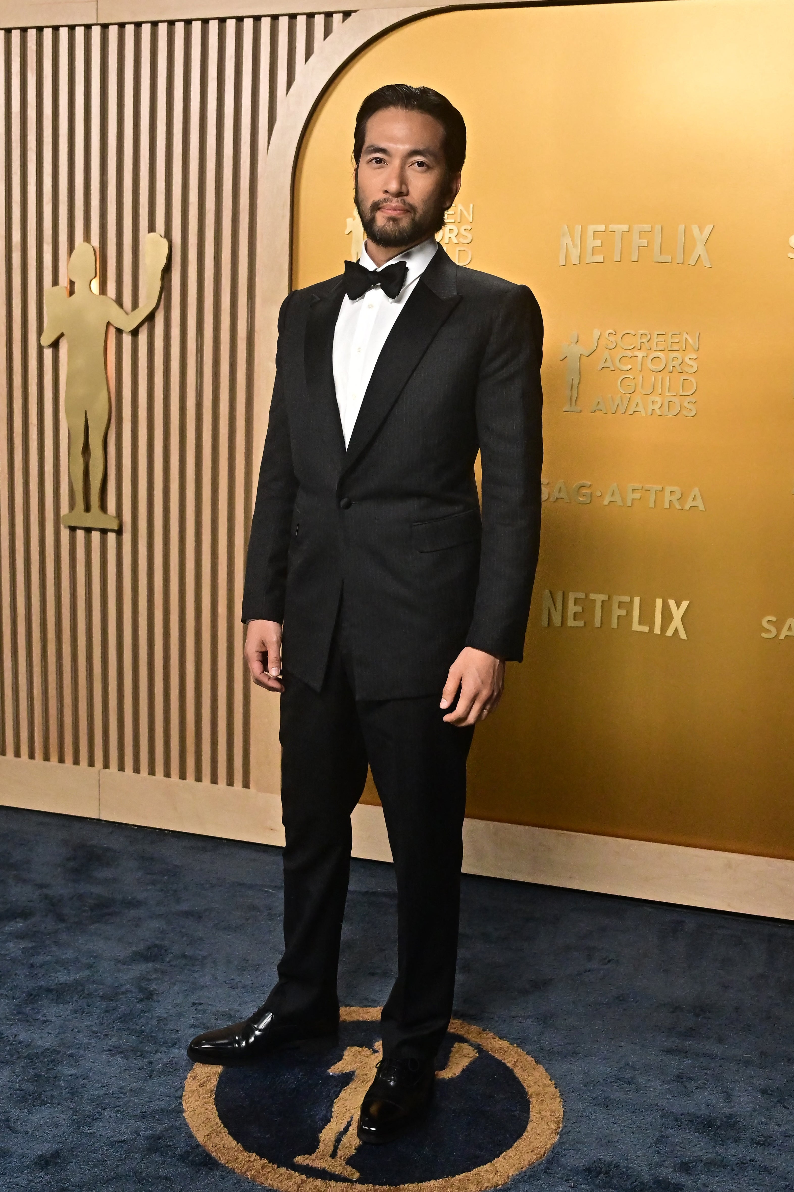 Person in a formal suit stands confidently on an industry event carpet with logo backdrop