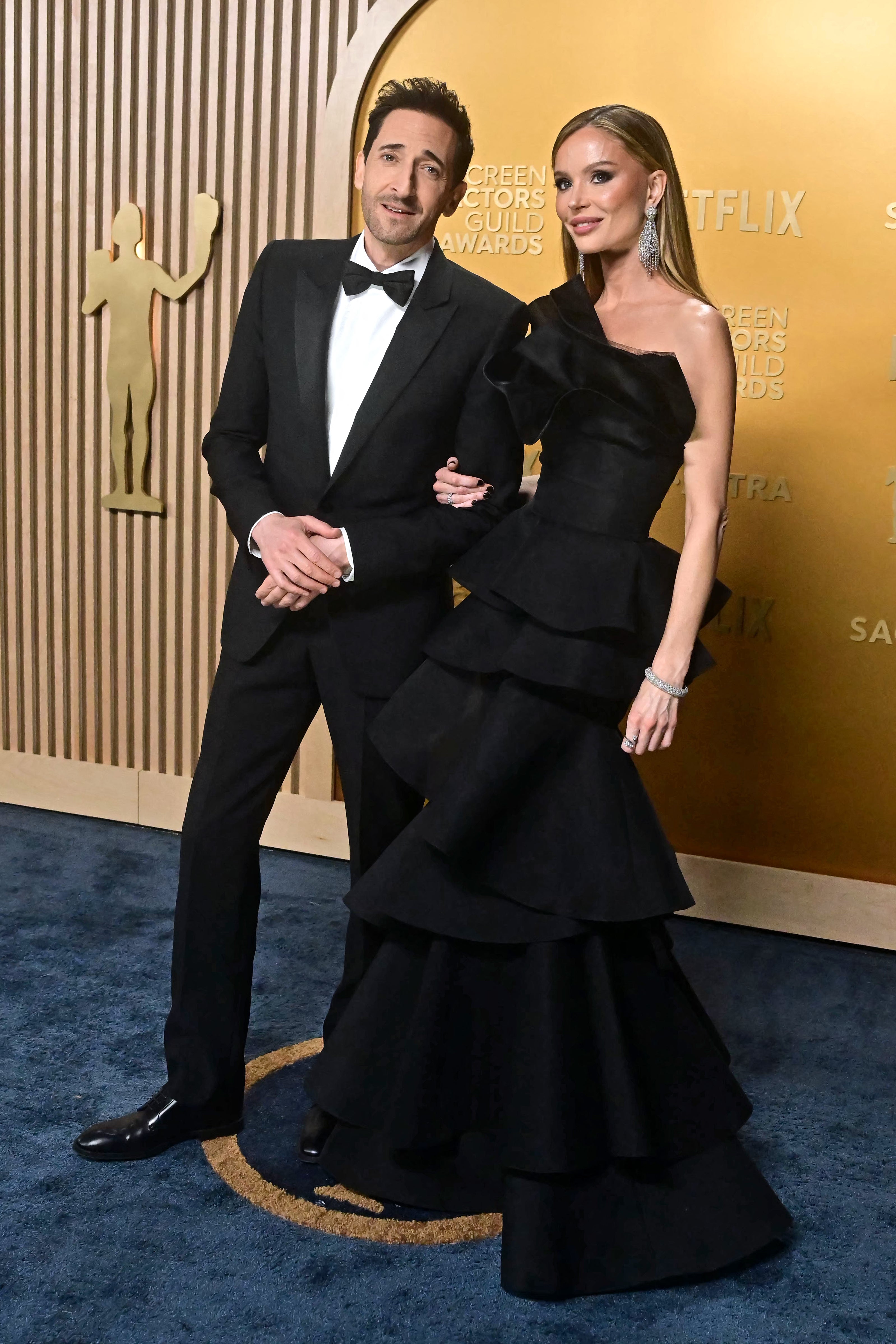 A man in a tuxedo and a woman in a tiered gown pose on the red carpet at an awards event