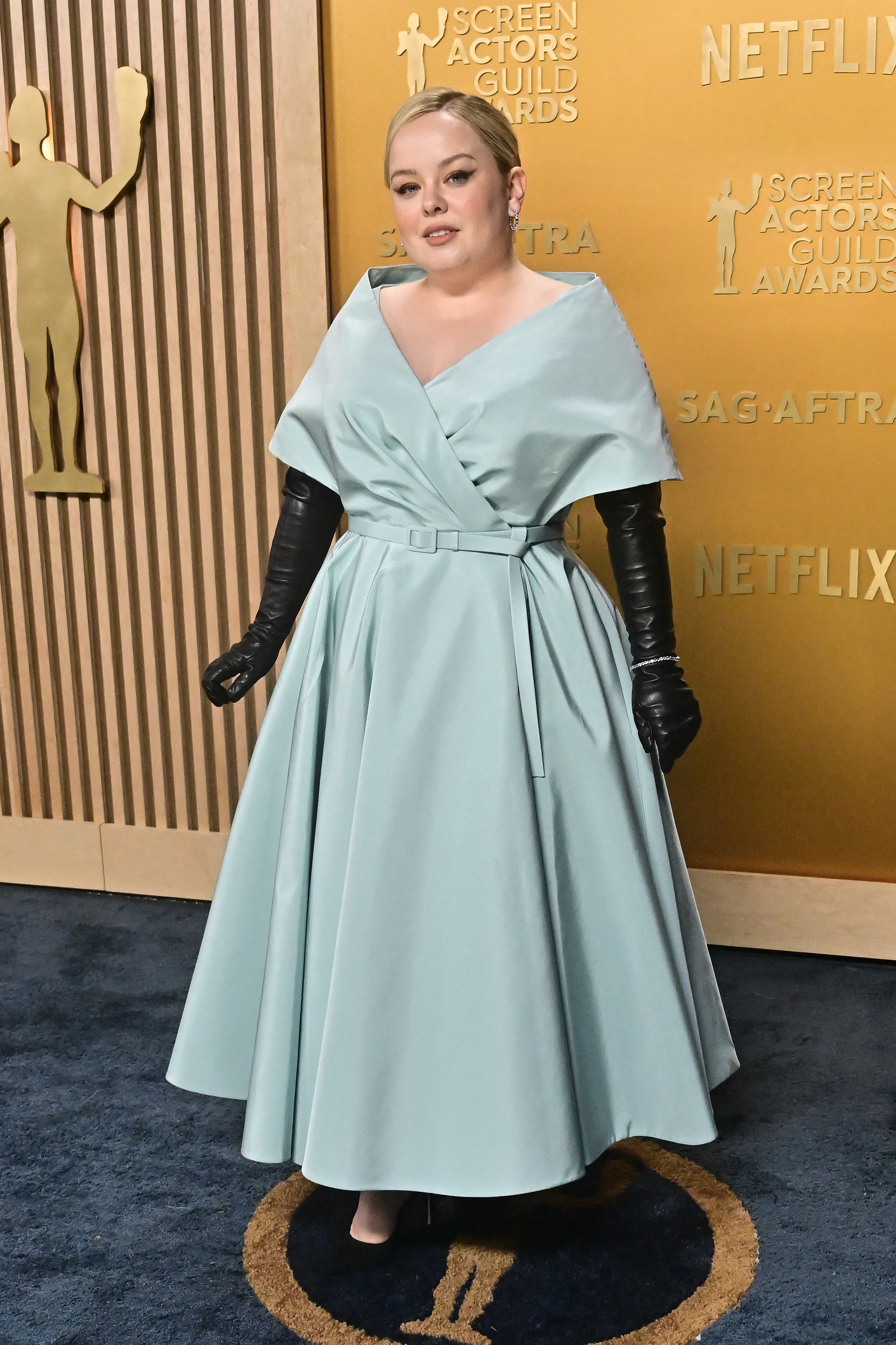 Person in formal gown with a wide skirt, cape-like top, and long black gloves at an awards event backdrop