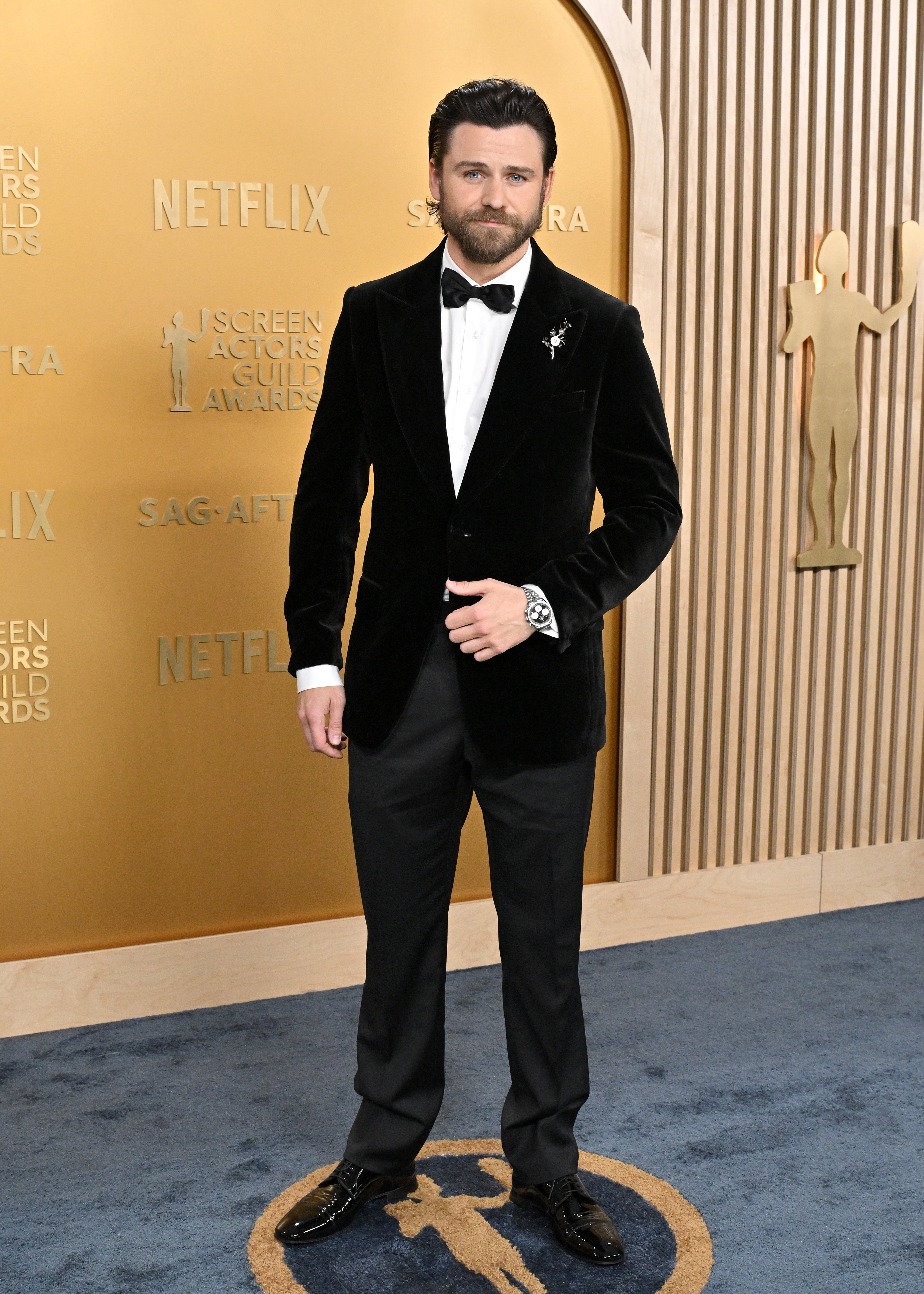 Person on red carpet in a tailored velvet suit, white shirt, and bow tie, standing in front of a backdrop with award show branding