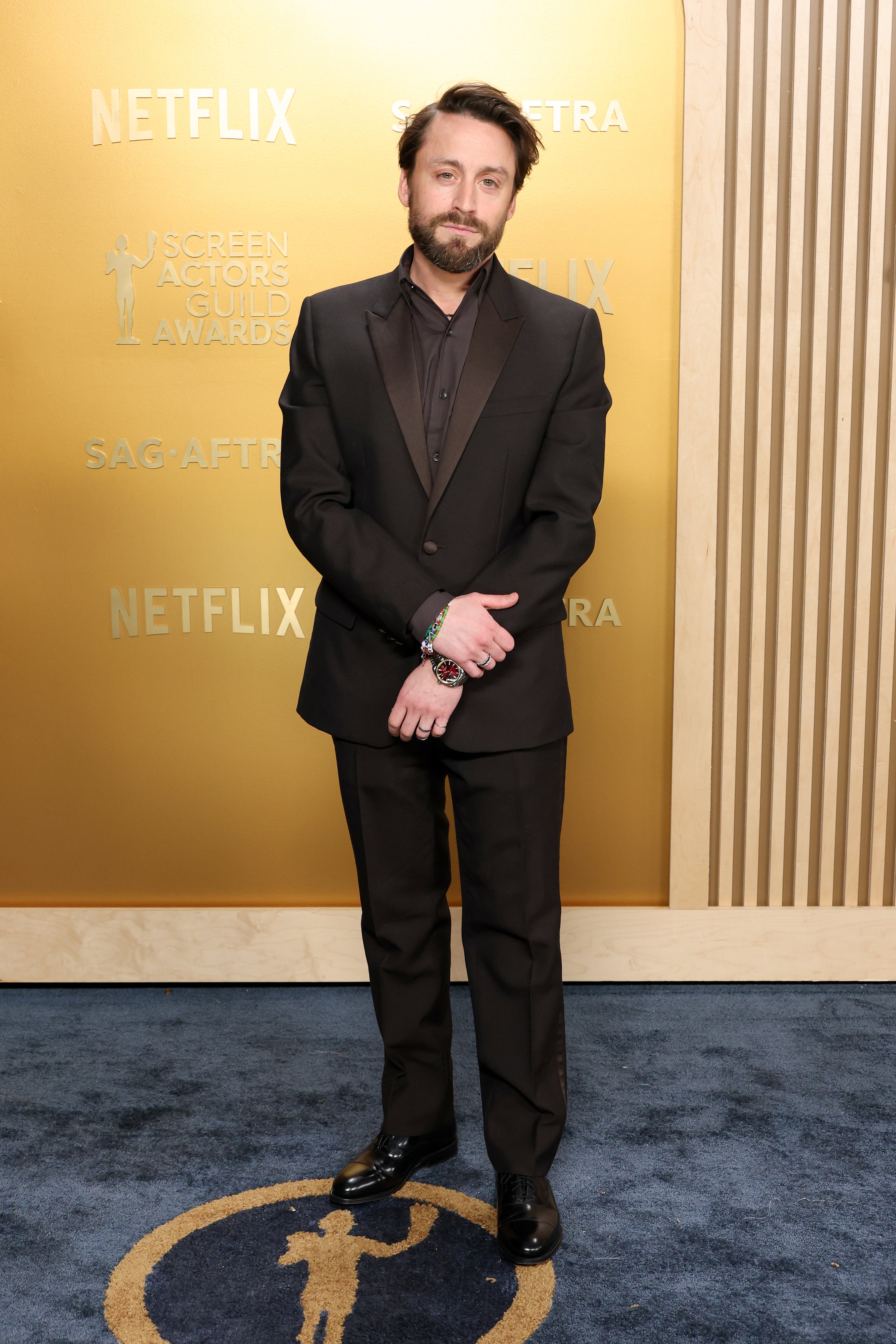Person on a red carpet in formal black suit and shiny shoes, standing in front of a backdrop with "Netflix" and awards logos