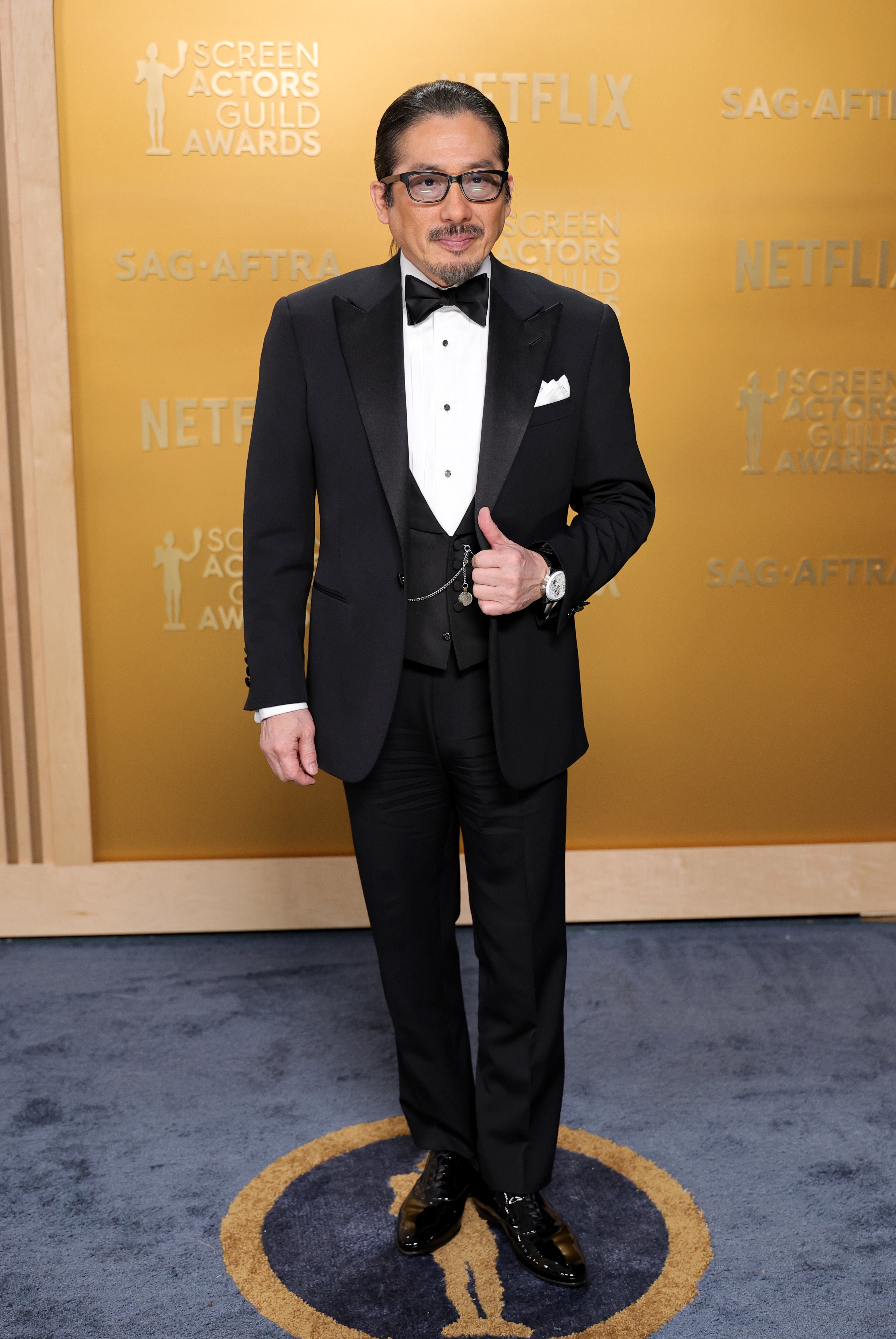 Person in a formal black tuxedo with a white shirt and bow tie, posing at an event with "Screen Actors Guild Awards" backdrop