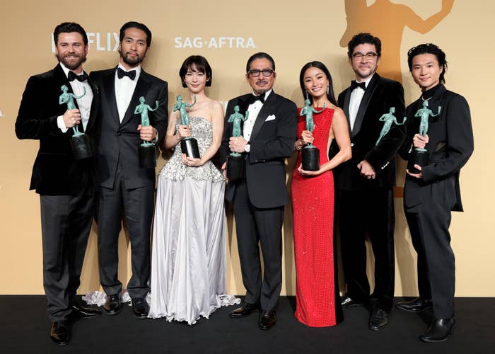 Group of actors in formal wear holding awards at a ceremony. Each person is smiling and posing for a photo