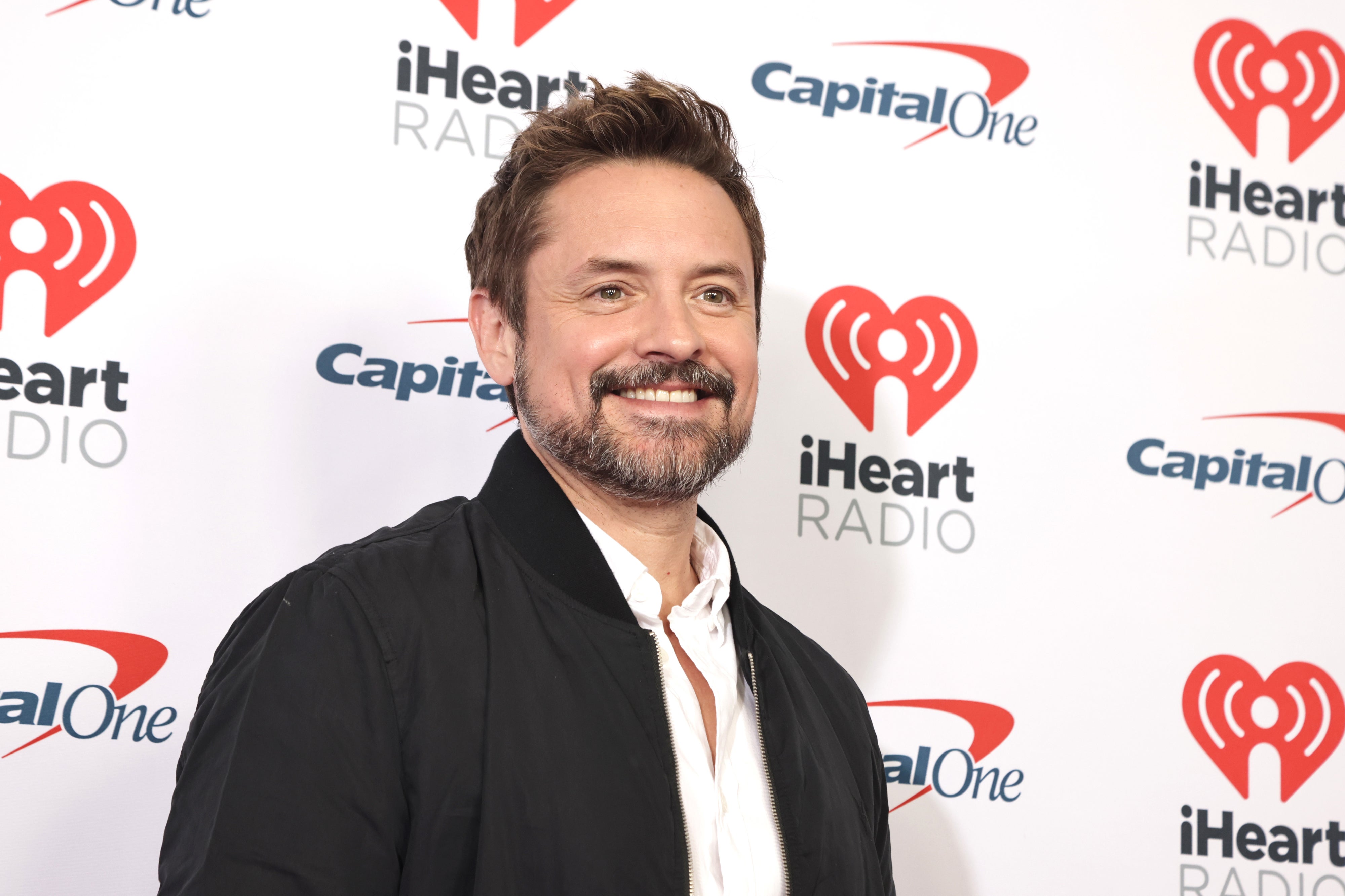Will Friedle in a jacket and shirt on the iHeartRadio red carpet event backdrop