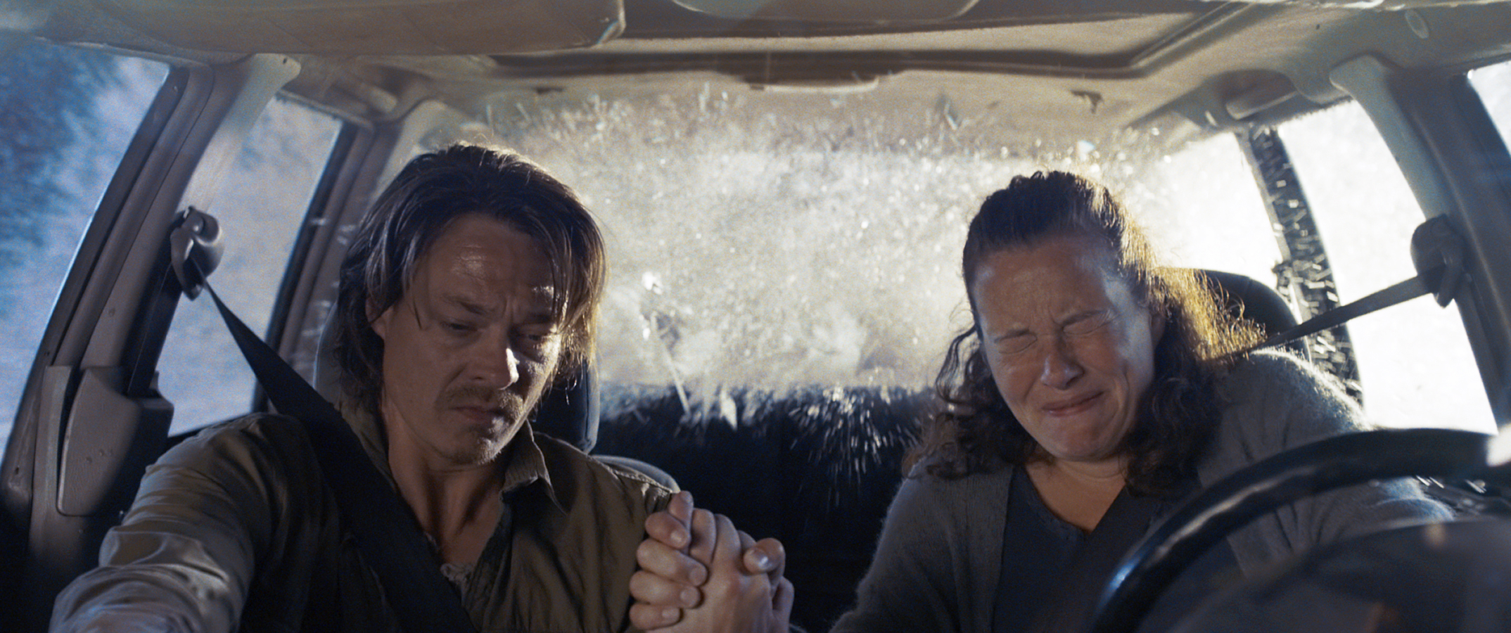 Two people in a car, visibly tense, holding hands as the windshield shatters and water floods in