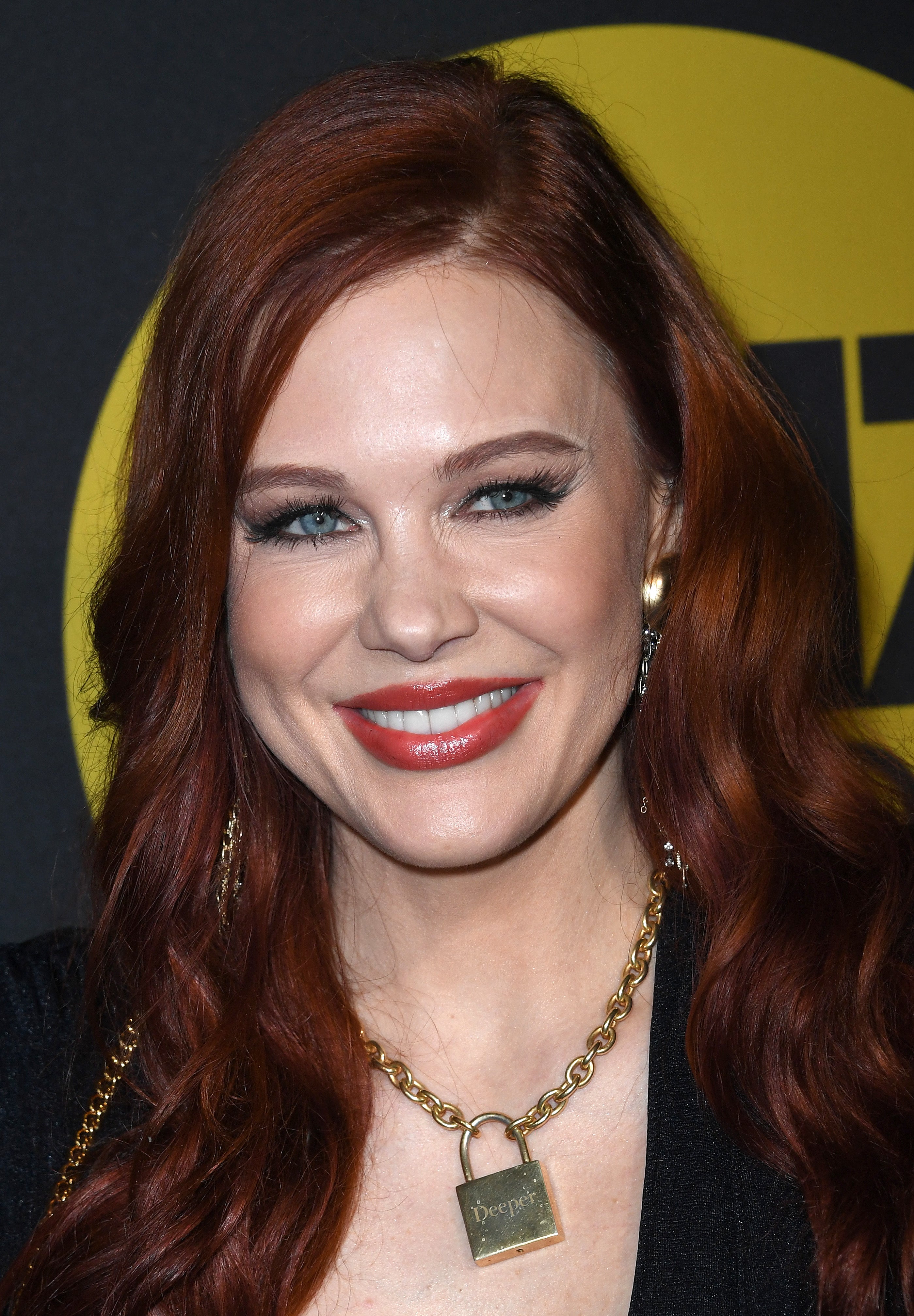 Maitland Ward with long hair and vibrant makeup smiles, wearing a black outfit and a chain necklace with a lock pendant