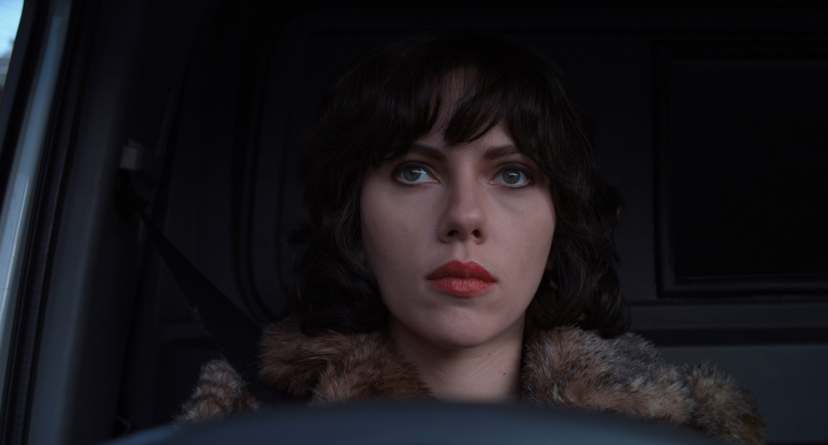 Scarlett Johansson wears short dark hair, red lipstick and a fur coat sitting in a vehicle, looking forward intently