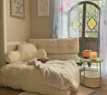 Cozy reading nook with a plush chair and pillows, a book on the seat, a small table with a candle, and decorative curtains in a bright room