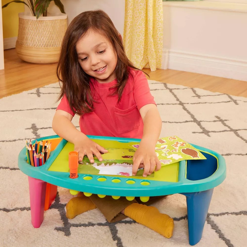 Anak yang duduk di atas tikar lantai, bermain dengan kerajinan di atas meja aktivitas kecil, dikelilingi oleh krayon dan stiker, tersenyum dan bertunangan