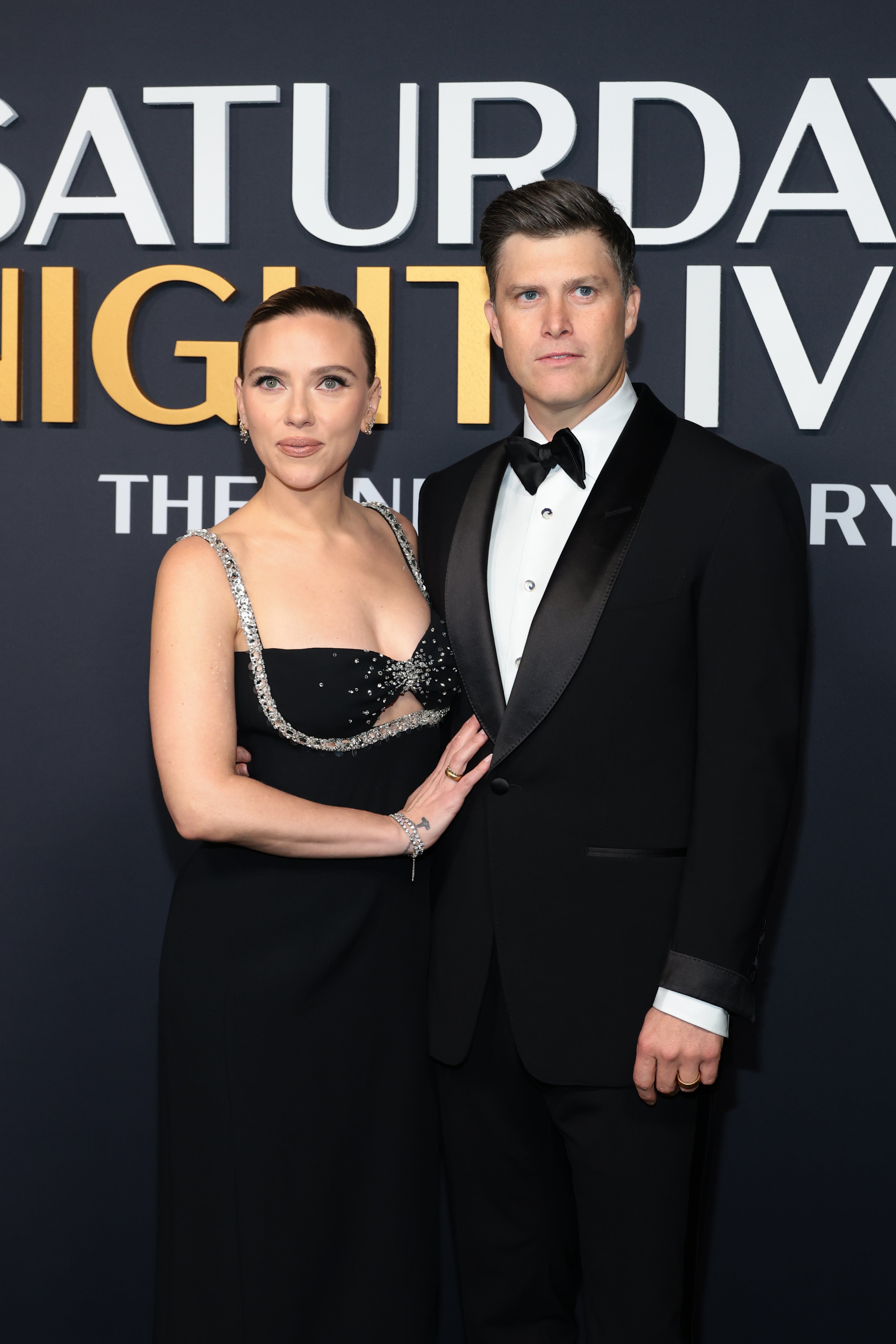 Two people on a red carpet; one in a black gown with silver details, the other in a classic black tuxedo, posing at a celebrity event