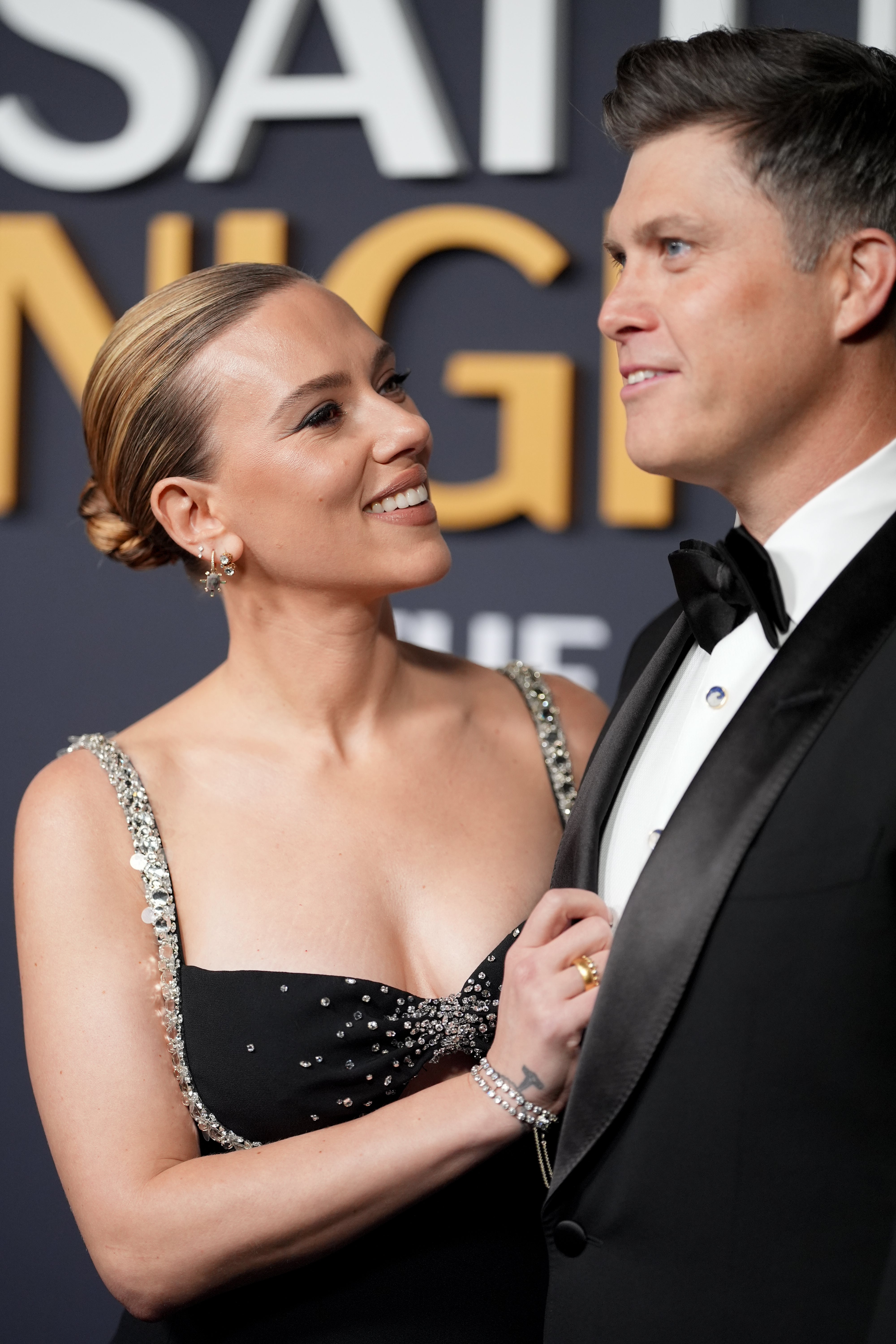 A woman in a chic black gown with sparkling straps smiles at a man in a classic tuxedo on a red carpet