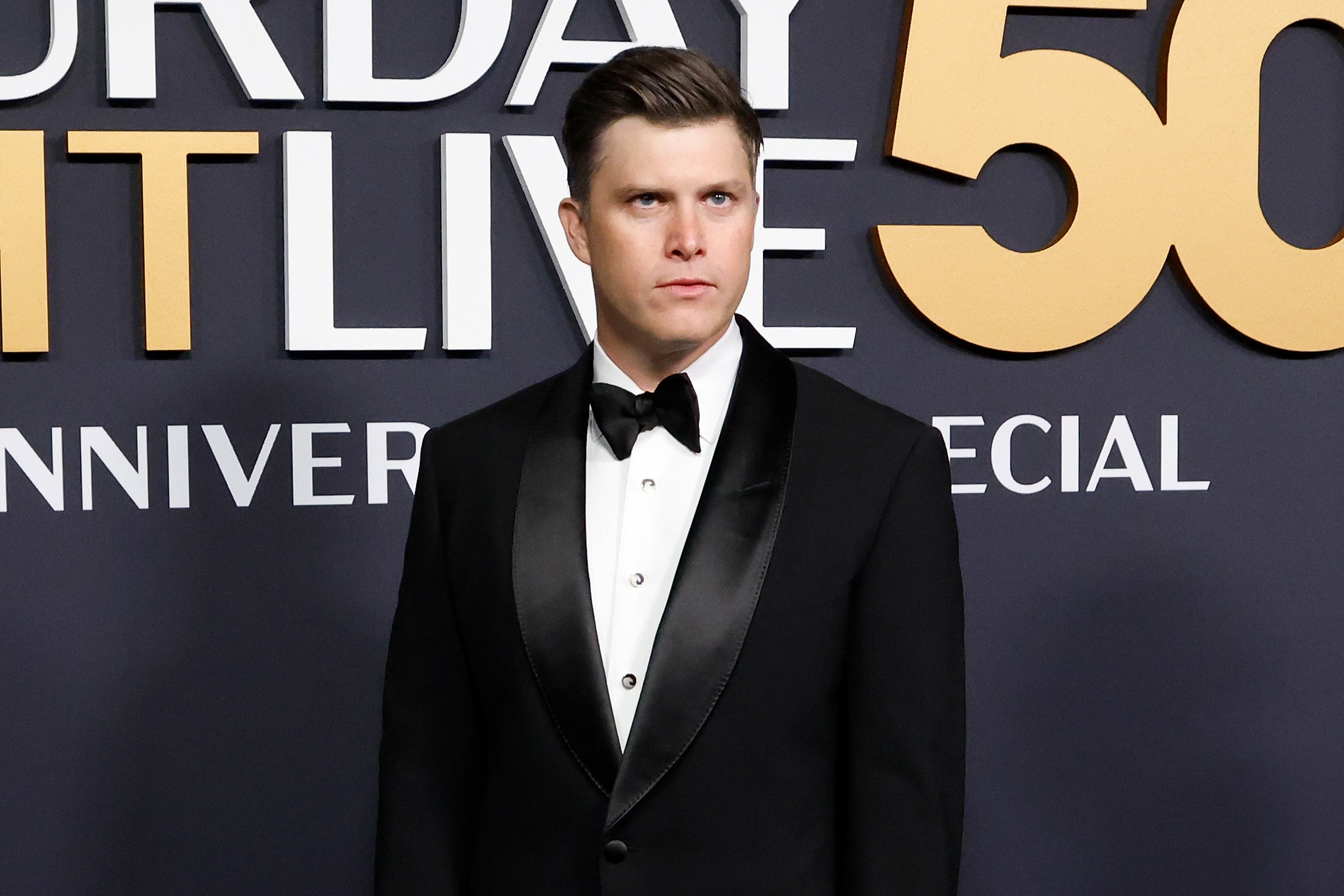 Man in black tuxedo stands in front of "Saturday Night Live 50th Anniversary Special" backdrop