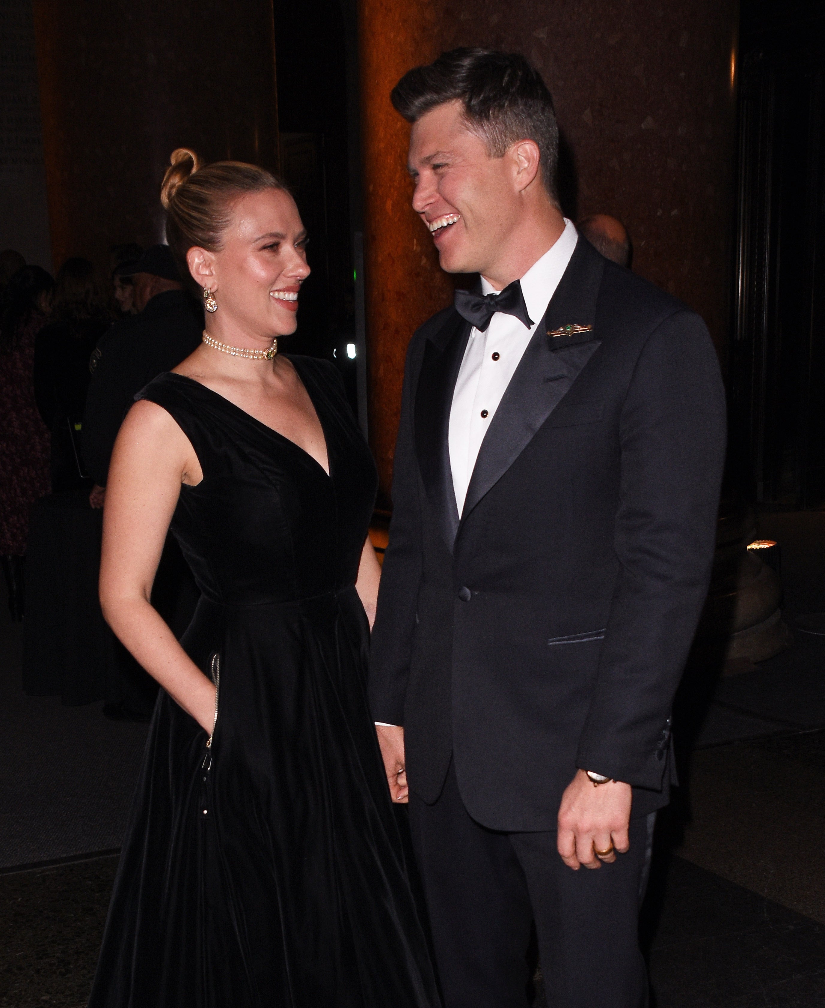 A couple on the red carpet, the woman in a sleeveless black gown and pearl necklace, the man in a classic tuxedo, both smiling at each other