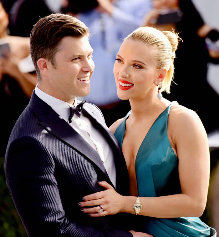A couple poses on the red carpet, with the woman in a stylish, sleeveless gown and the man in a tuxedo, smiling at each other