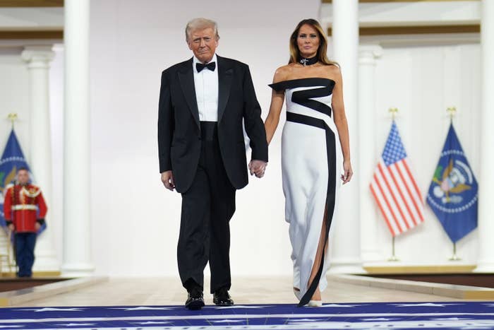 A man in a tuxedo and a woman in a stylish off-the-shoulder gown walk hand in hand on a formal event stage, with U.S. flags in the background