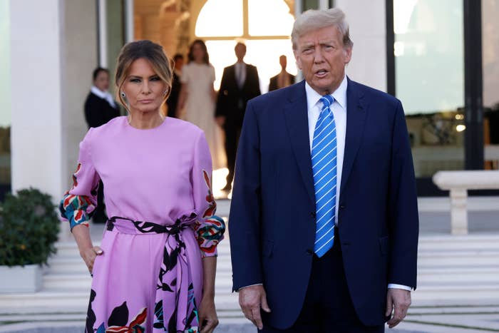 A man in a suit and tie stands beside a woman in a floral-patterned dress outside a building, with people visible in the background