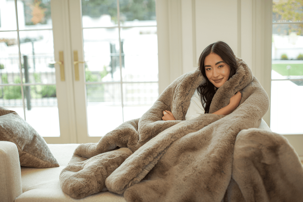 Model wrapped in plush light brown blanket, sitting on a couch by a large window, looking comfortable and relaxed