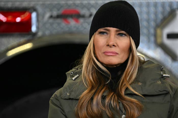 A woman in a dark coat and beanie appears serious, standing in front of a backdrop with metal and machinery