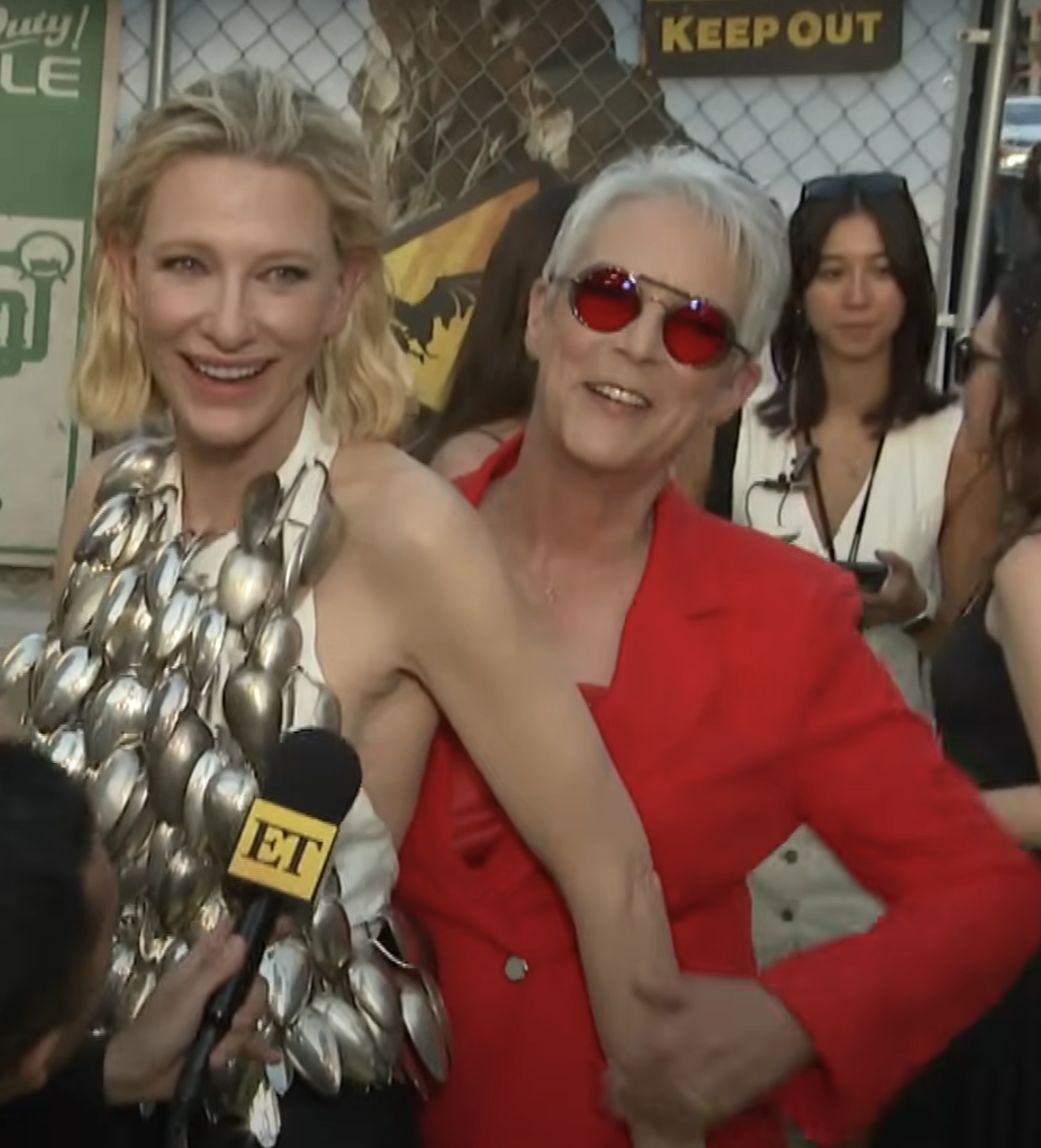 Cate in a unique silver outfit and Jamie in a suit with sunglasses, pose with Jamie behind Cate on a red carpet with an "ET" microphone visible