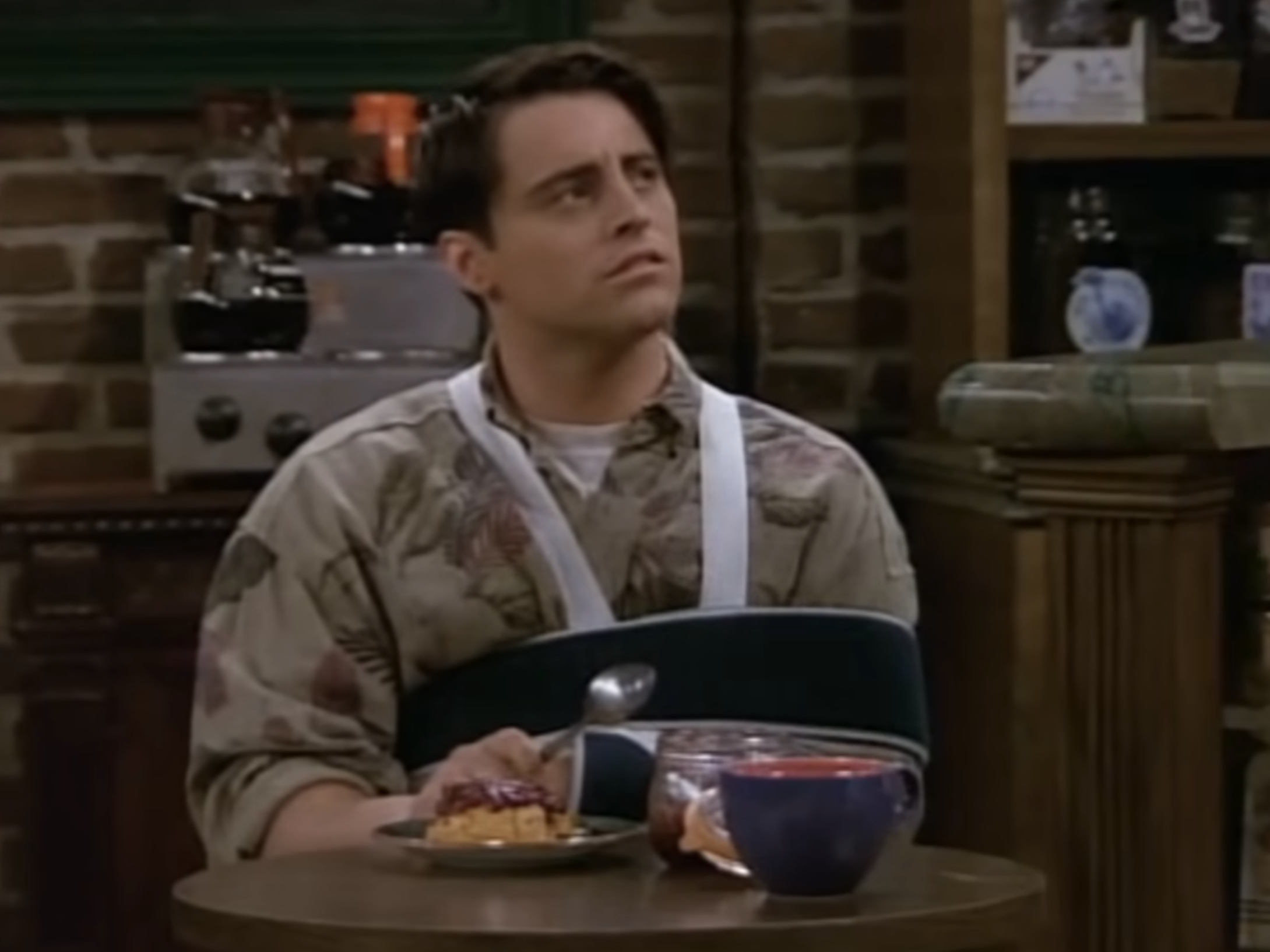 Person with arm in a sling, sitting at a café table, holding a spoon, looking up thoughtfully. Pie and a drink are on the table