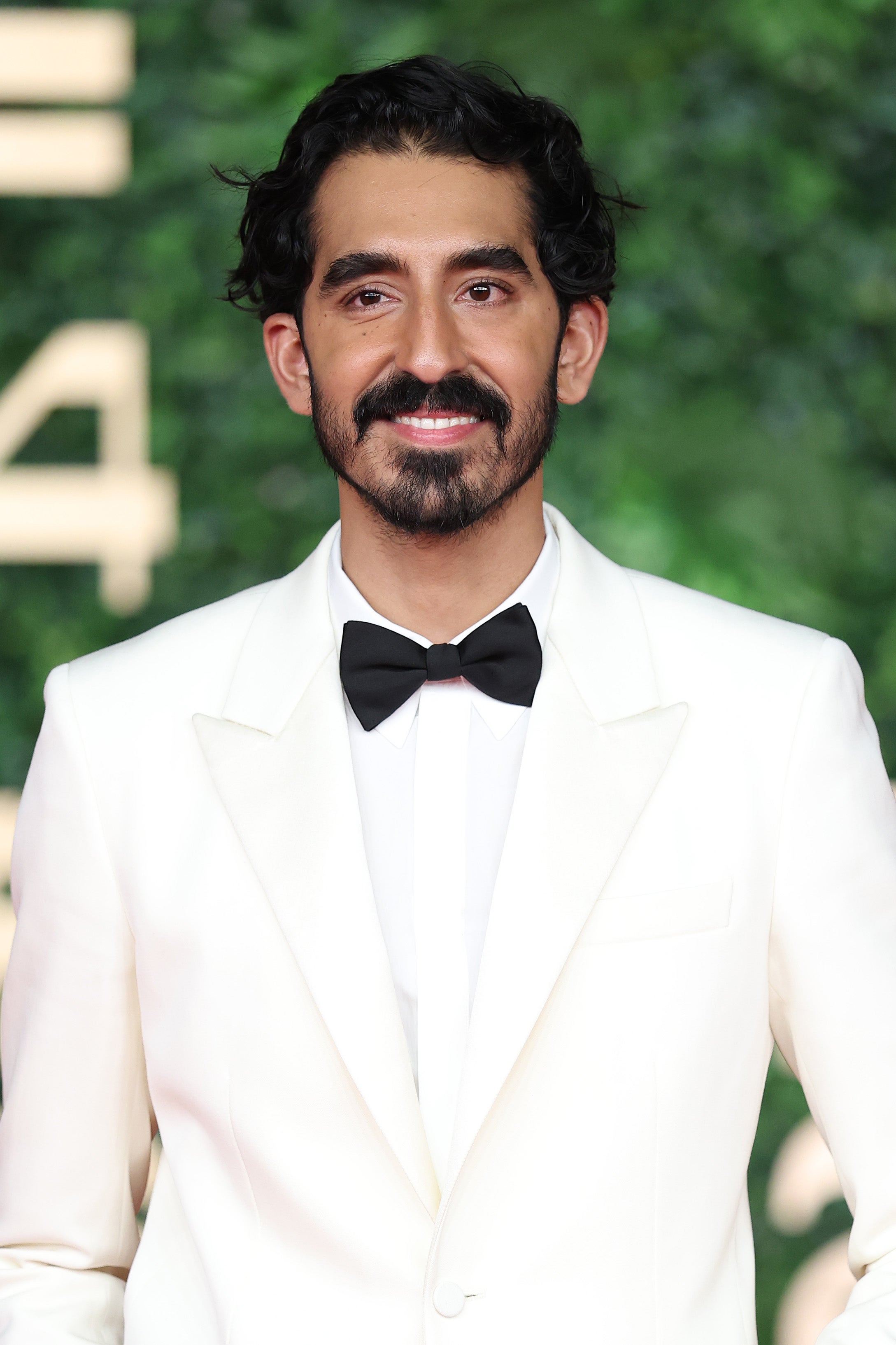 Person in a formal white tuxedo with a black bow tie, standing in front of a greenery backdrop