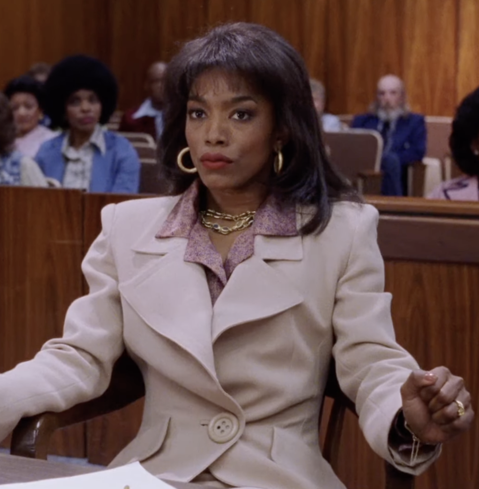 Person in a courtroom setting, wearing a stylish beige blazer with gold accessories, seated seriously at a desk