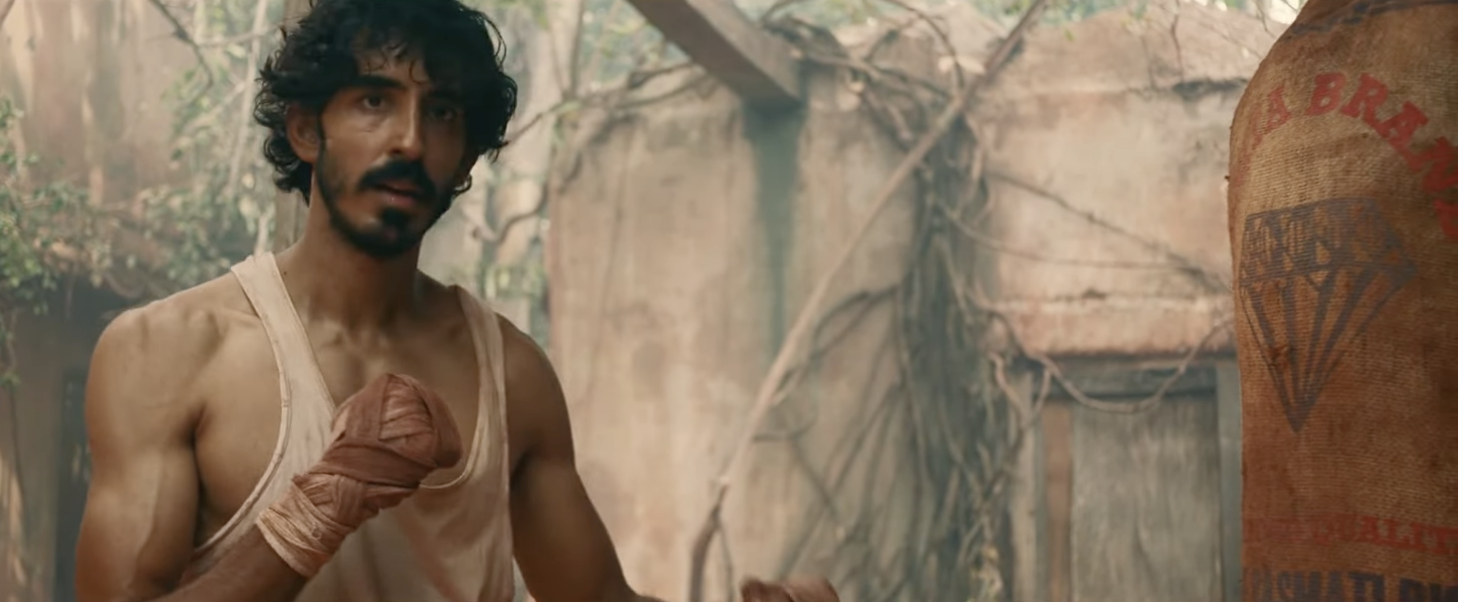 A person in a torn tank top and boxing hand wraps is practicing punches on a hanging punching bag in a rustic setting