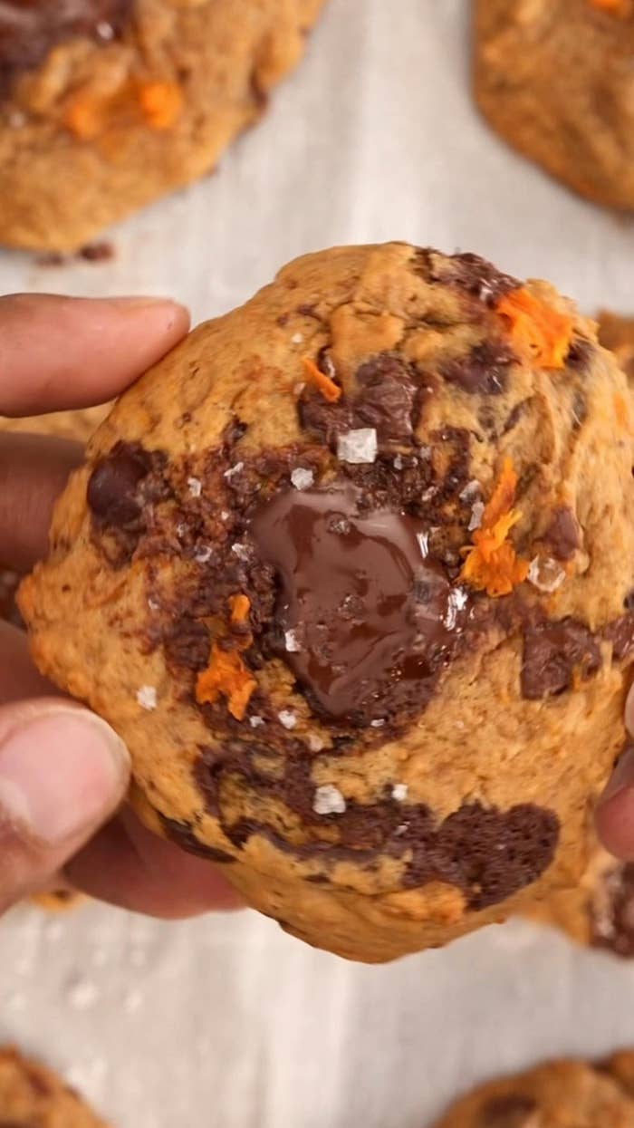 A hand holding a freshly baked cookie with melted chocolate and bits of orange topping visible