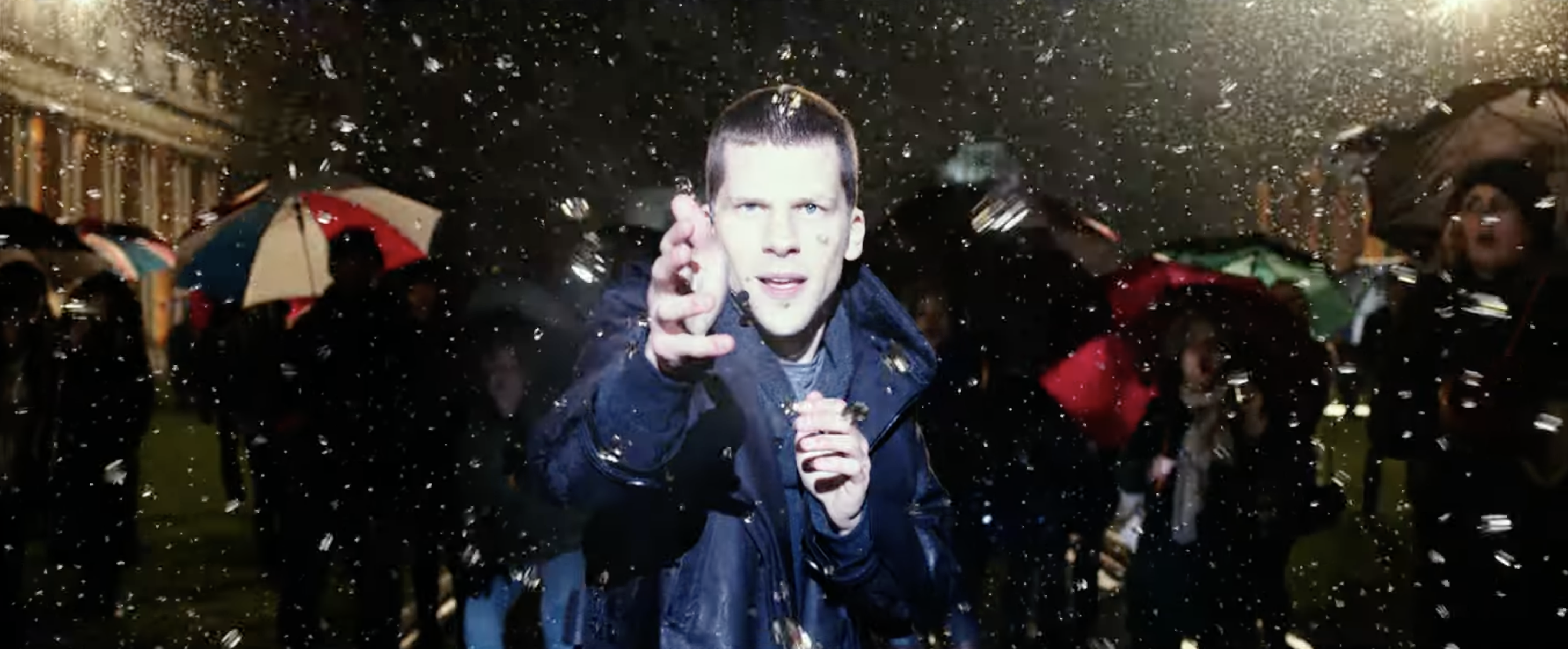 Person in a dark coat reaching towards the camera amidst falling confetti, surrounded by people with umbrellas at an outdoor event